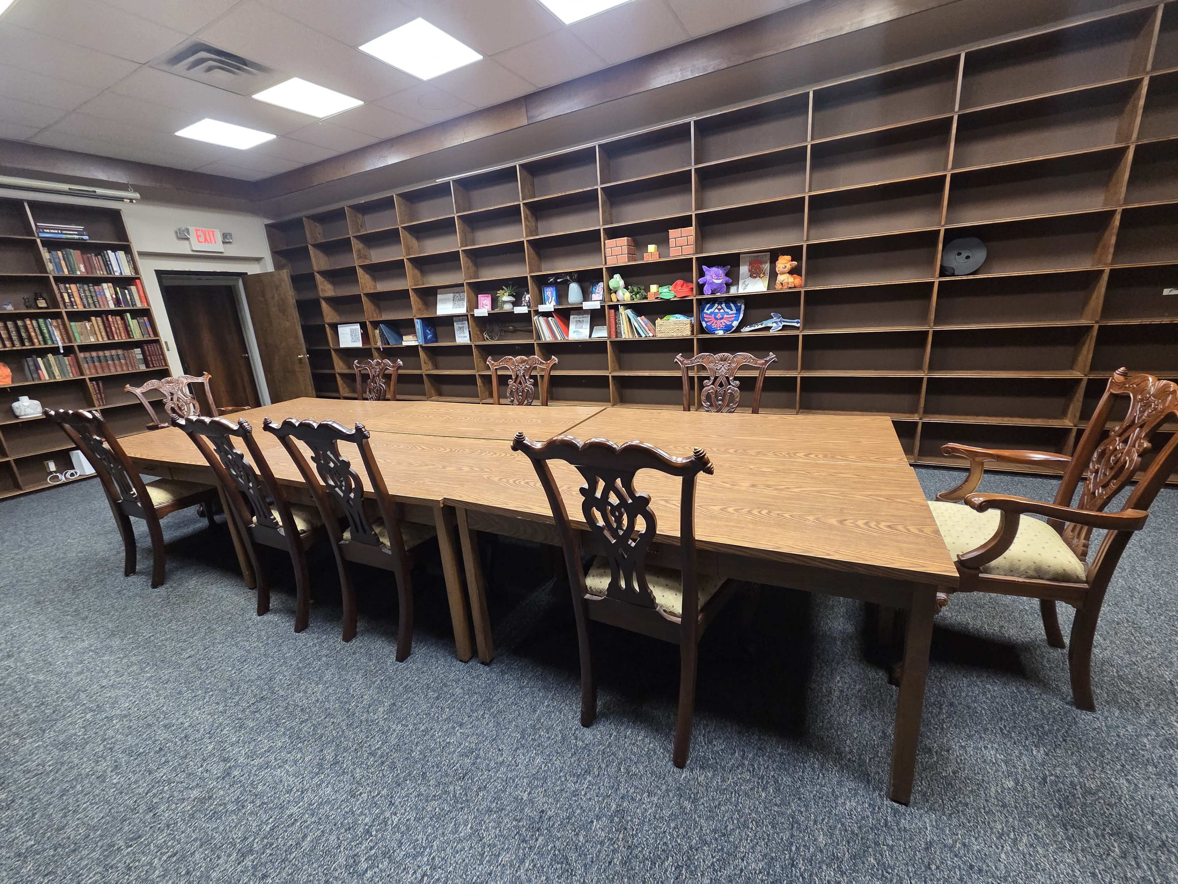 The image shows a large wooden conference table surrounded by intricately designed chairs in a room with bookshelves filled with various books.