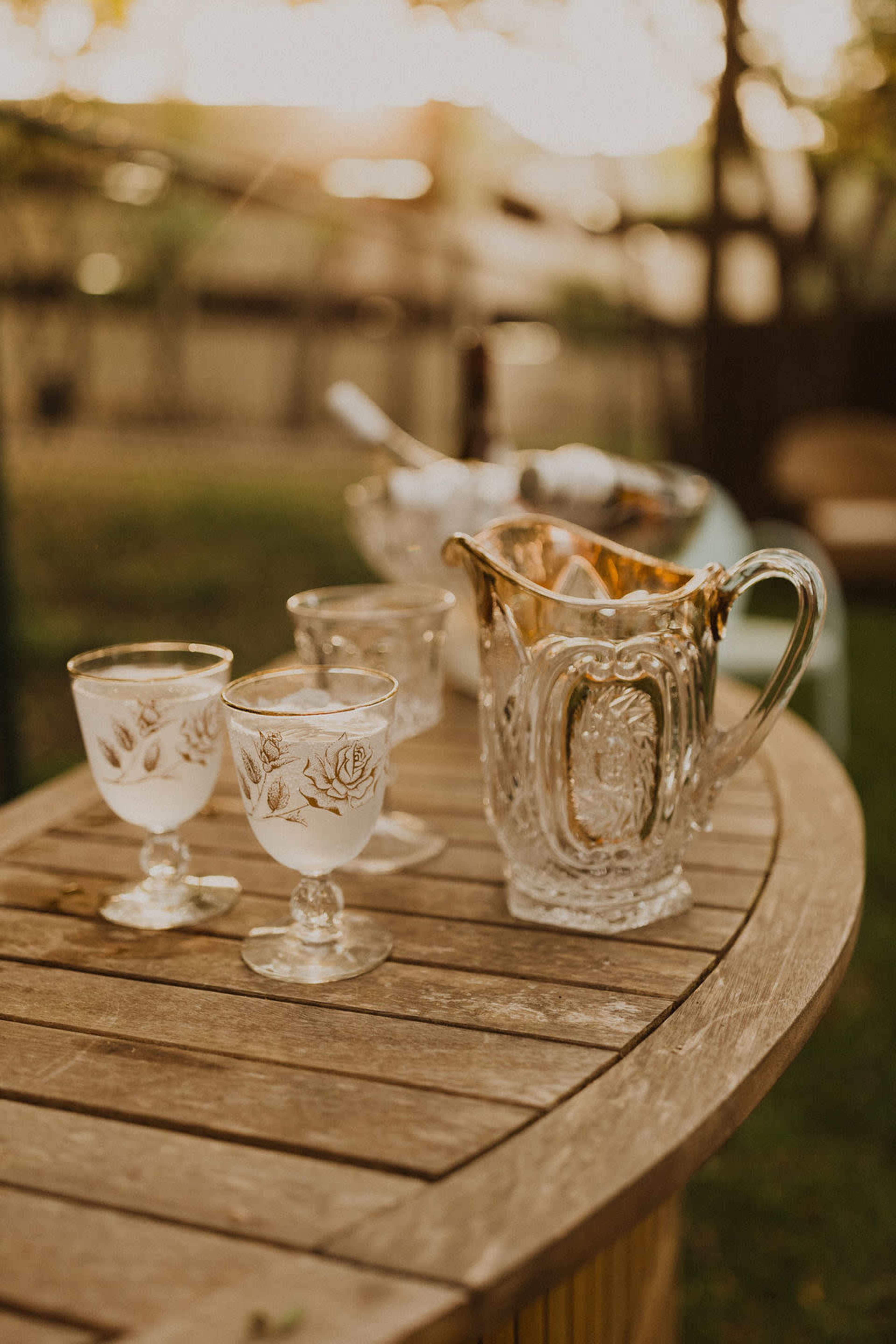 A wooden table displays a set of ornate glass cups and a decorative glass pitcher in a garden setting.