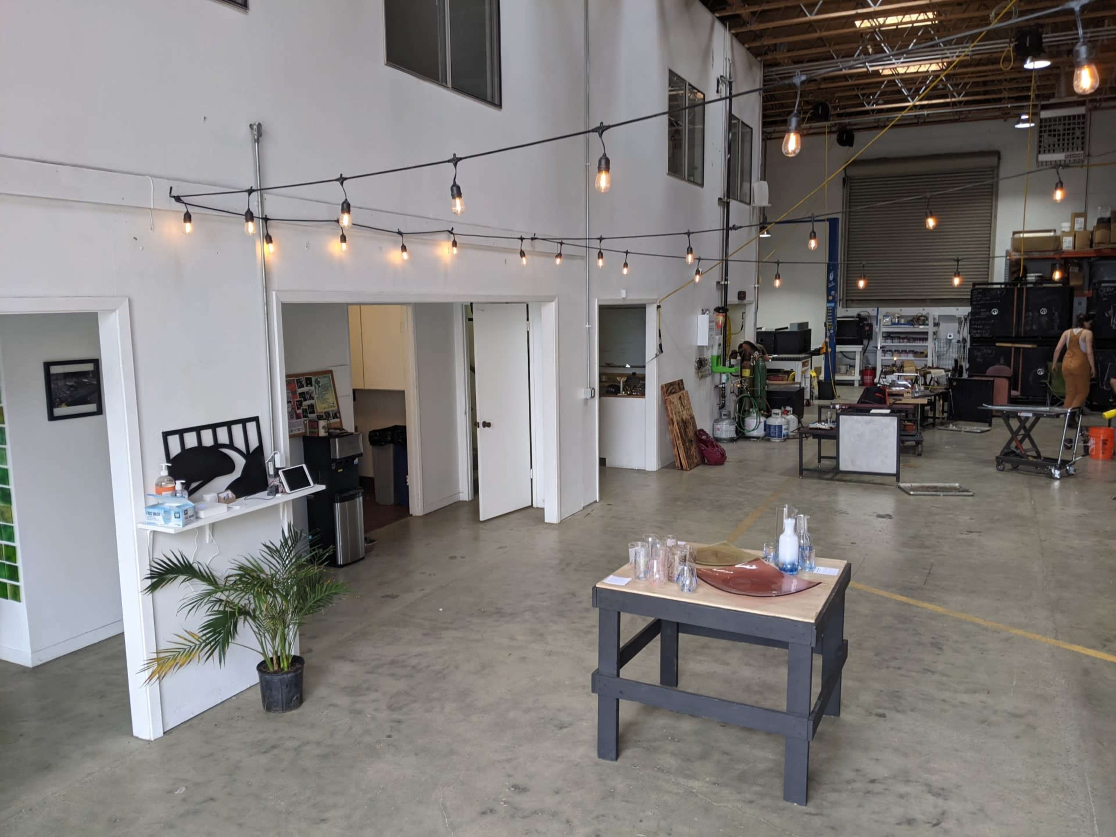 A spacious industrial-style room with a central table, string lights overhead, and various clutter and work areas visible in the background.