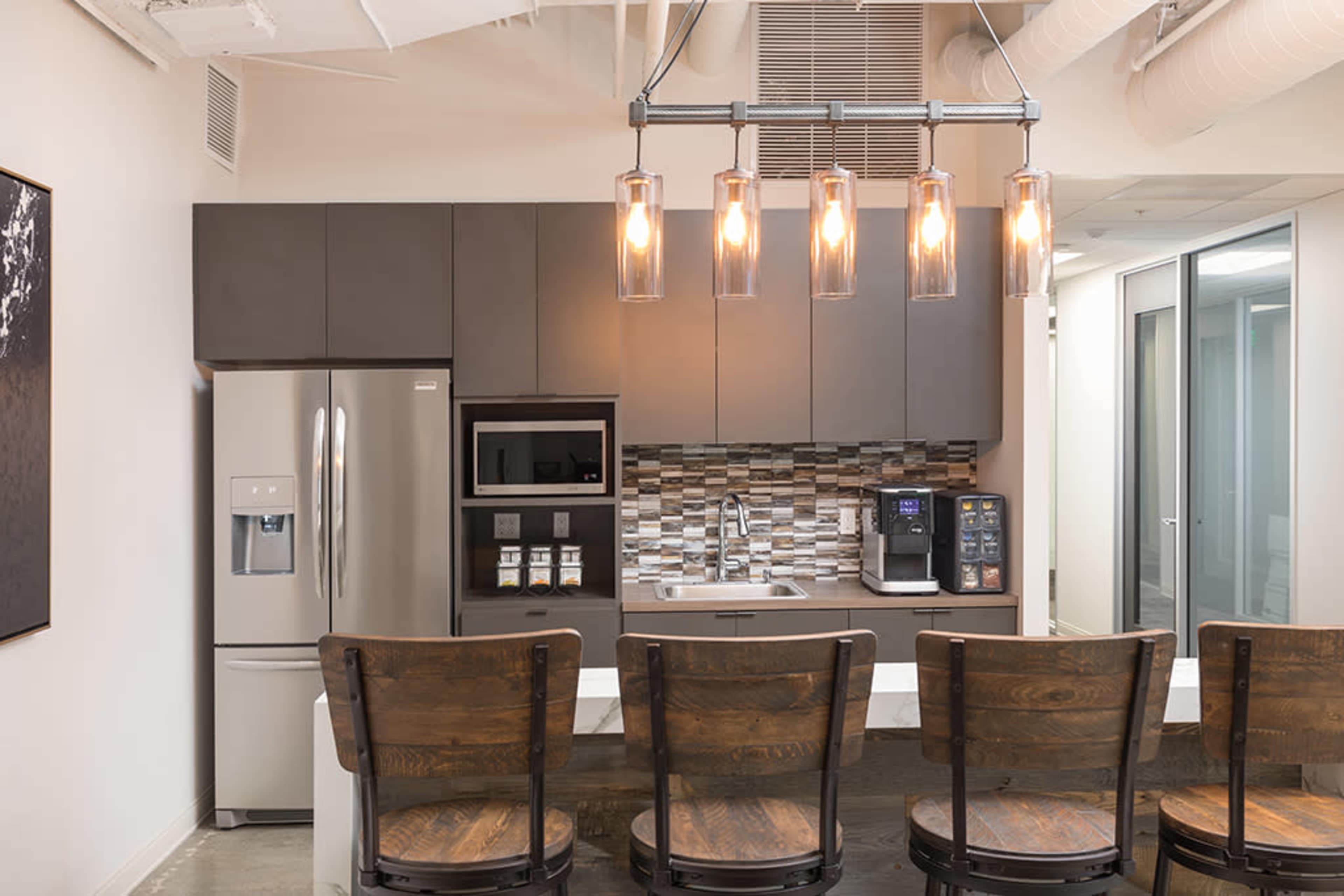 A modern kitchen features a refrigerator, oven, and coffee maker, with a bar counter and wooden chairs below hanging pendant lights.