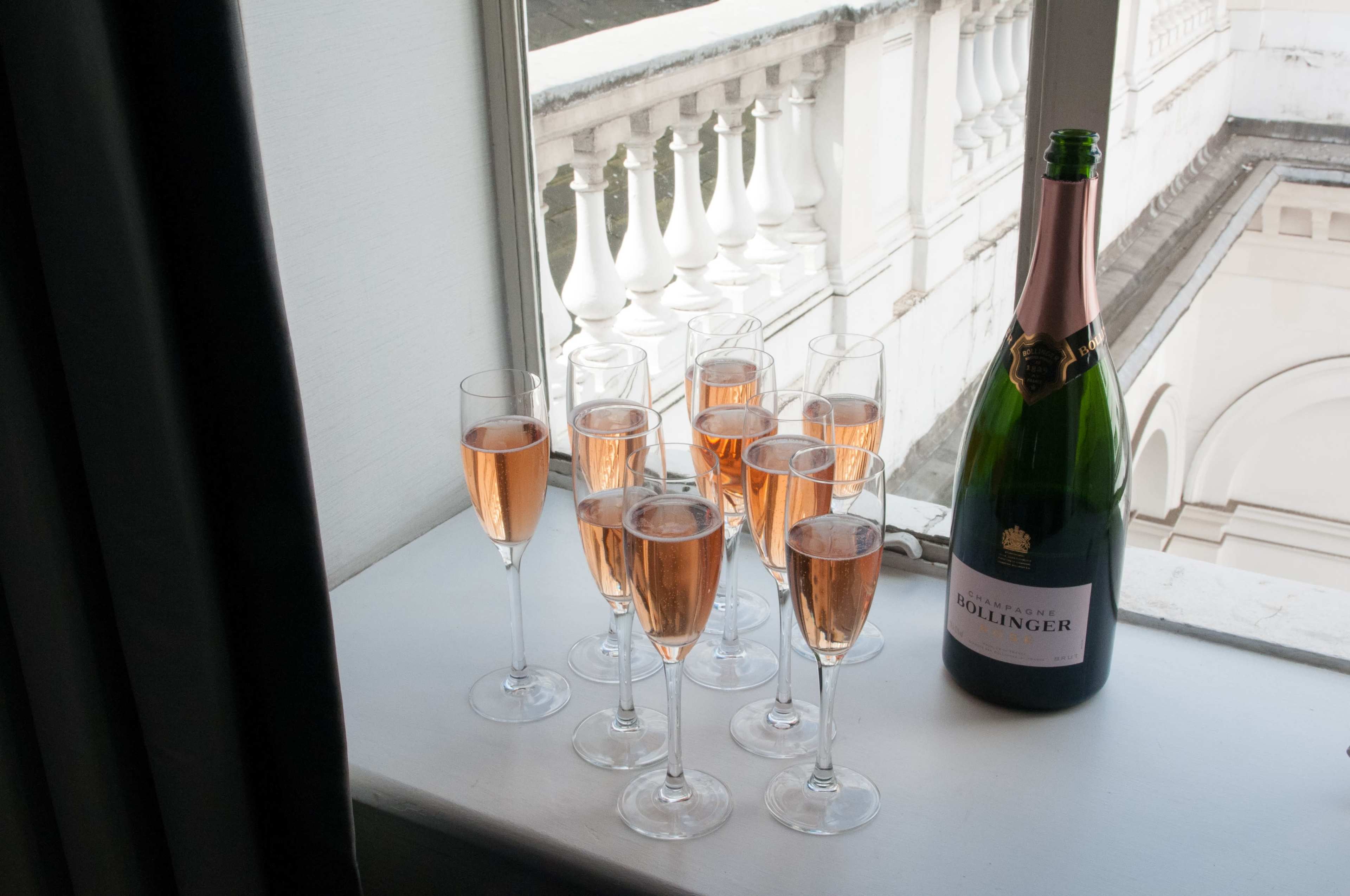 A bottle of Bollinger champagne sits beside nine glasses filled with sparkling rosé, placed on a windowsill.