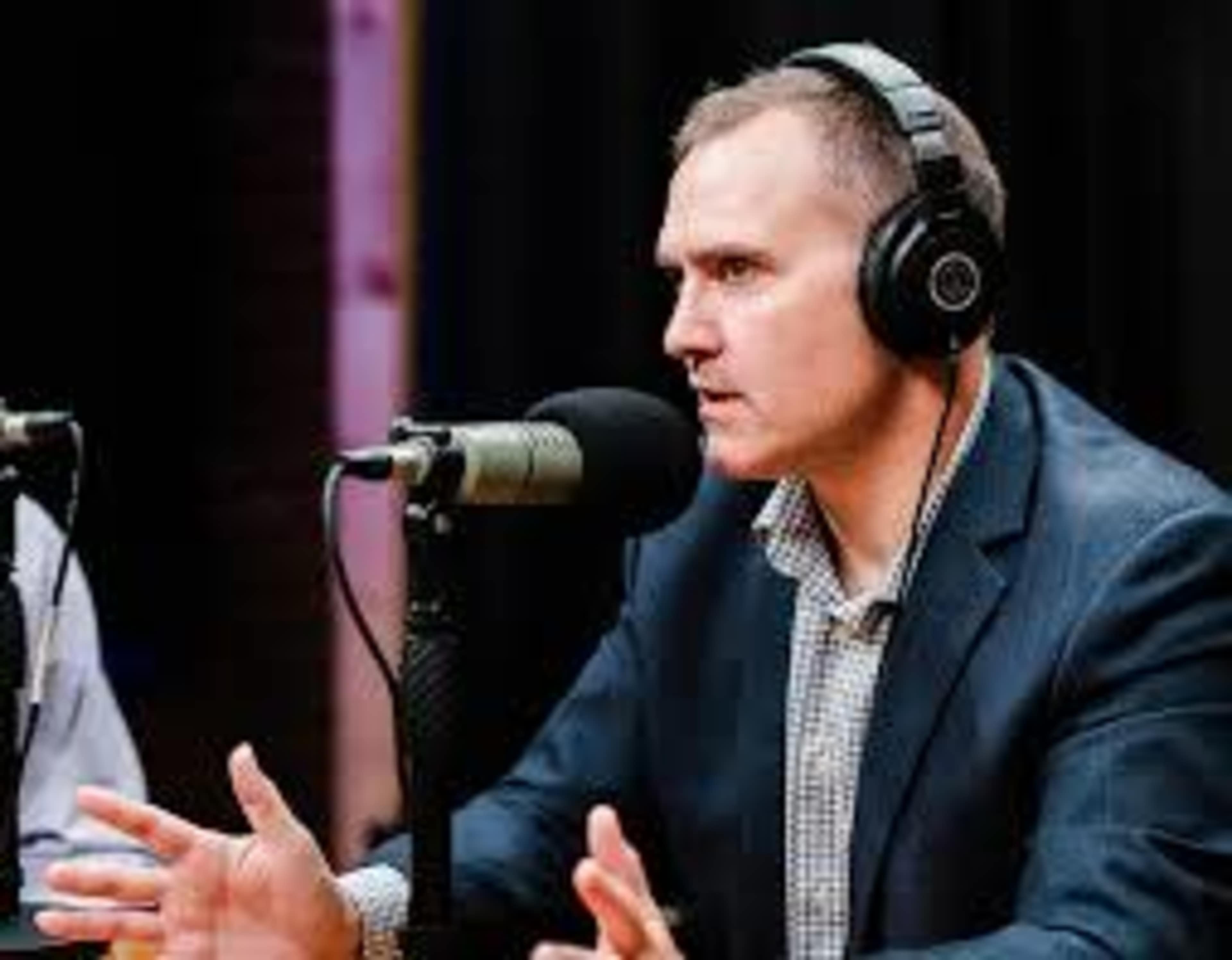 A man in a suit is speaking into a microphone while wearing headphones in a podcast recording setting.