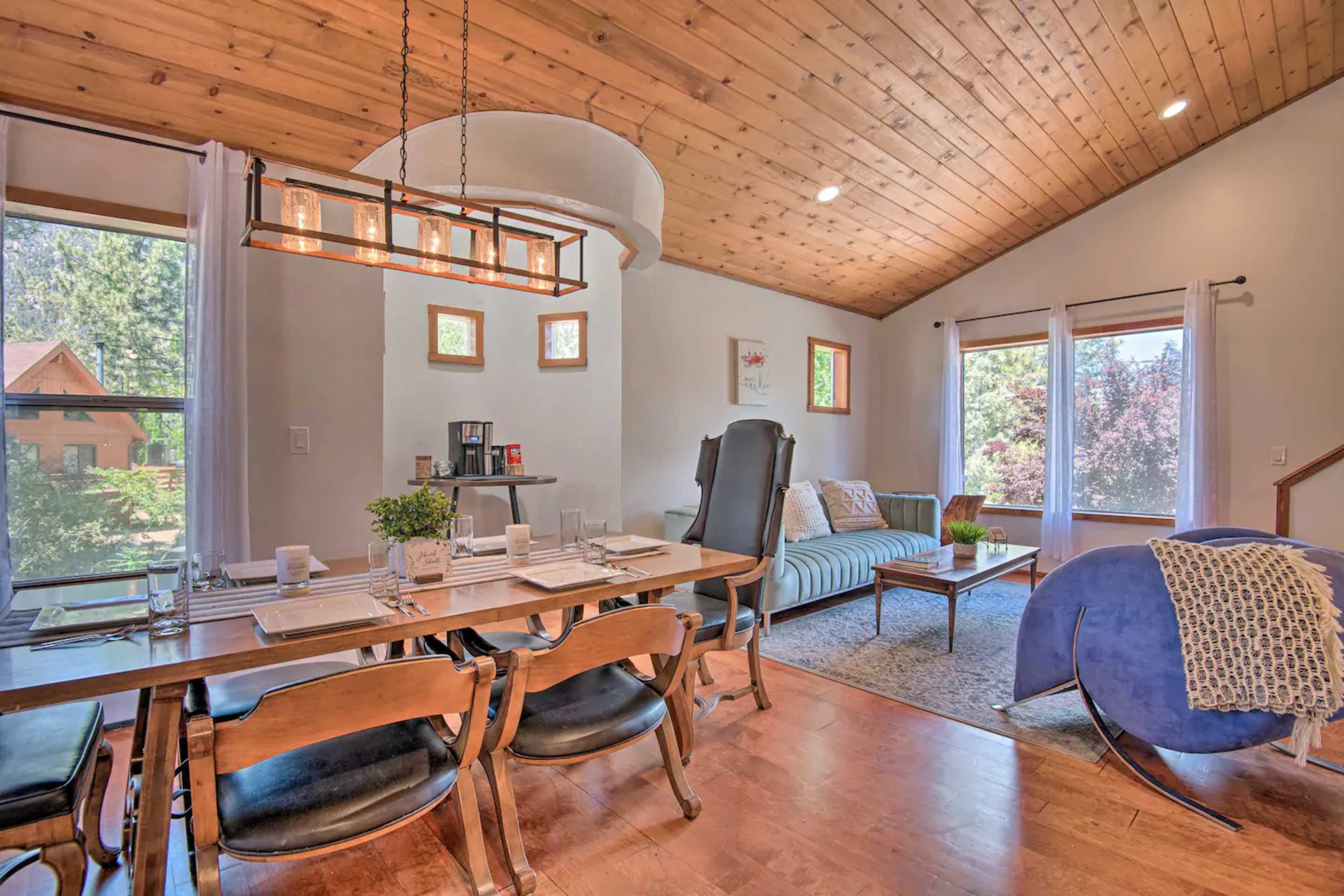 A dining area with a wooden table set for a meal, adjacent to a living room featuring a sofa and large windows that allow natural light to fill the space.