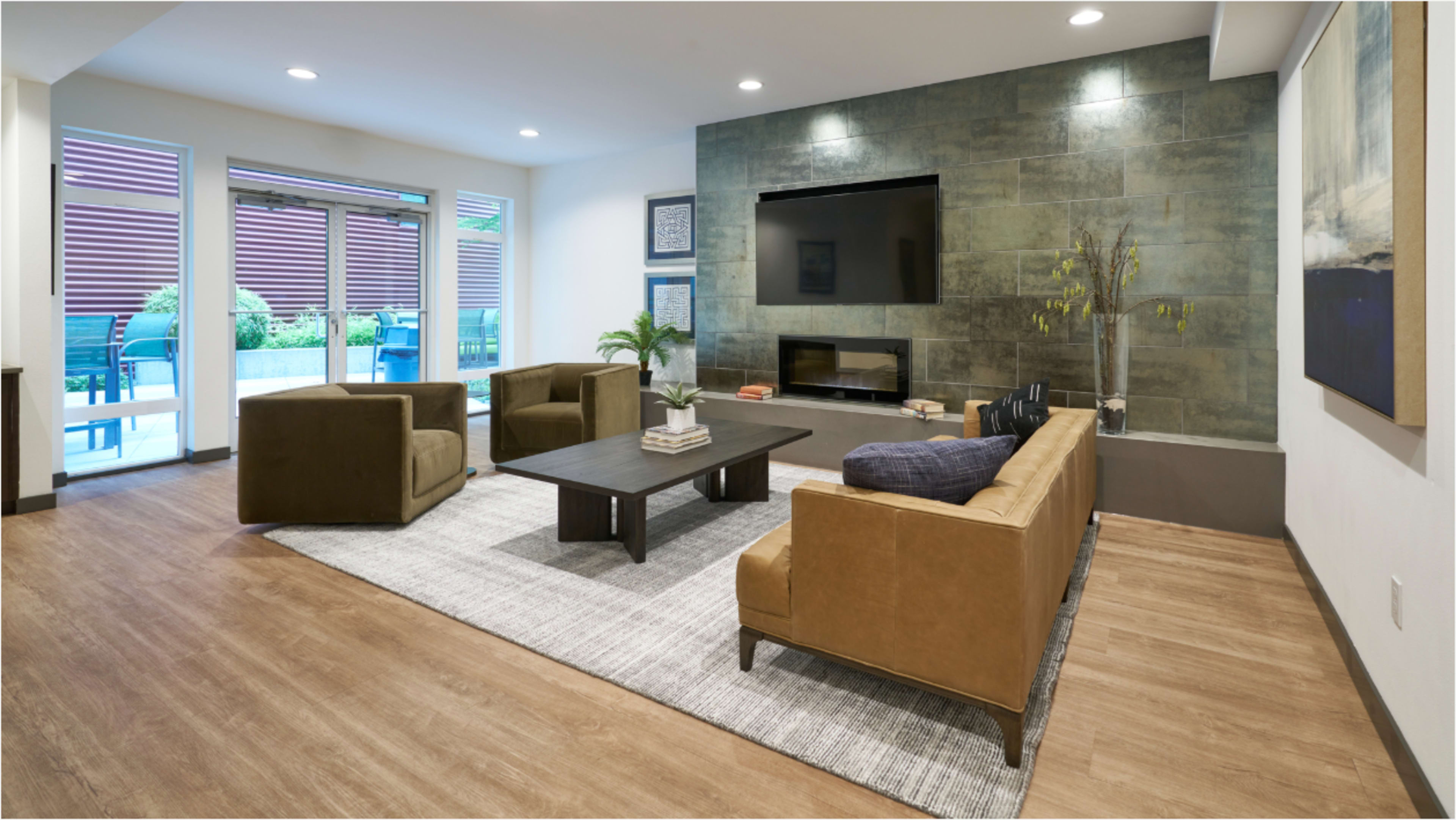 The image shows a modern living room featuring two sofas, a coffee table, and a large television mounted on a stone wall, with sliding glass doors leading to an outdoor area.