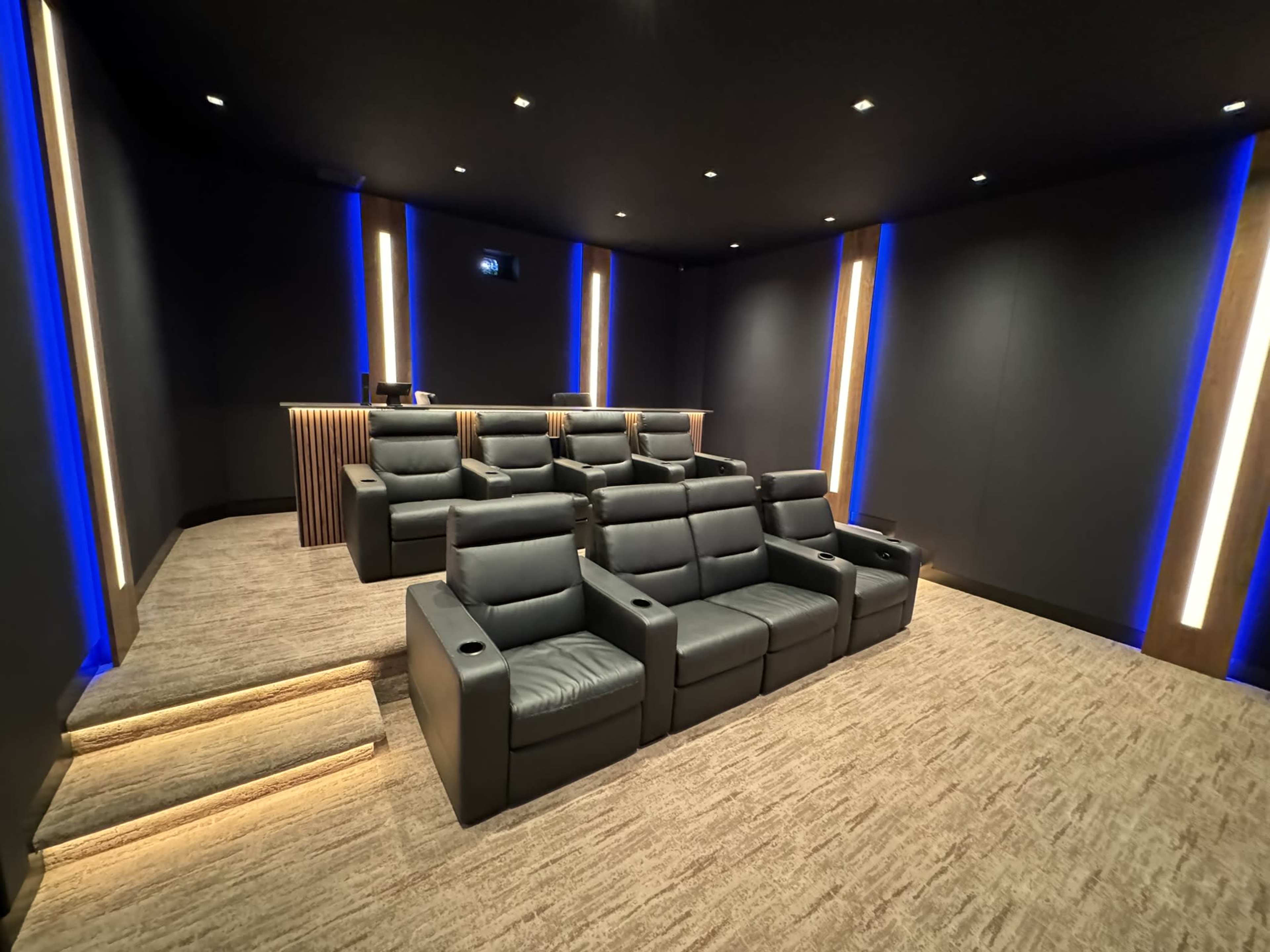 A modern home theater with black reclining chairs arranged in a tiered layout against a dark wall with blue accent lighting.