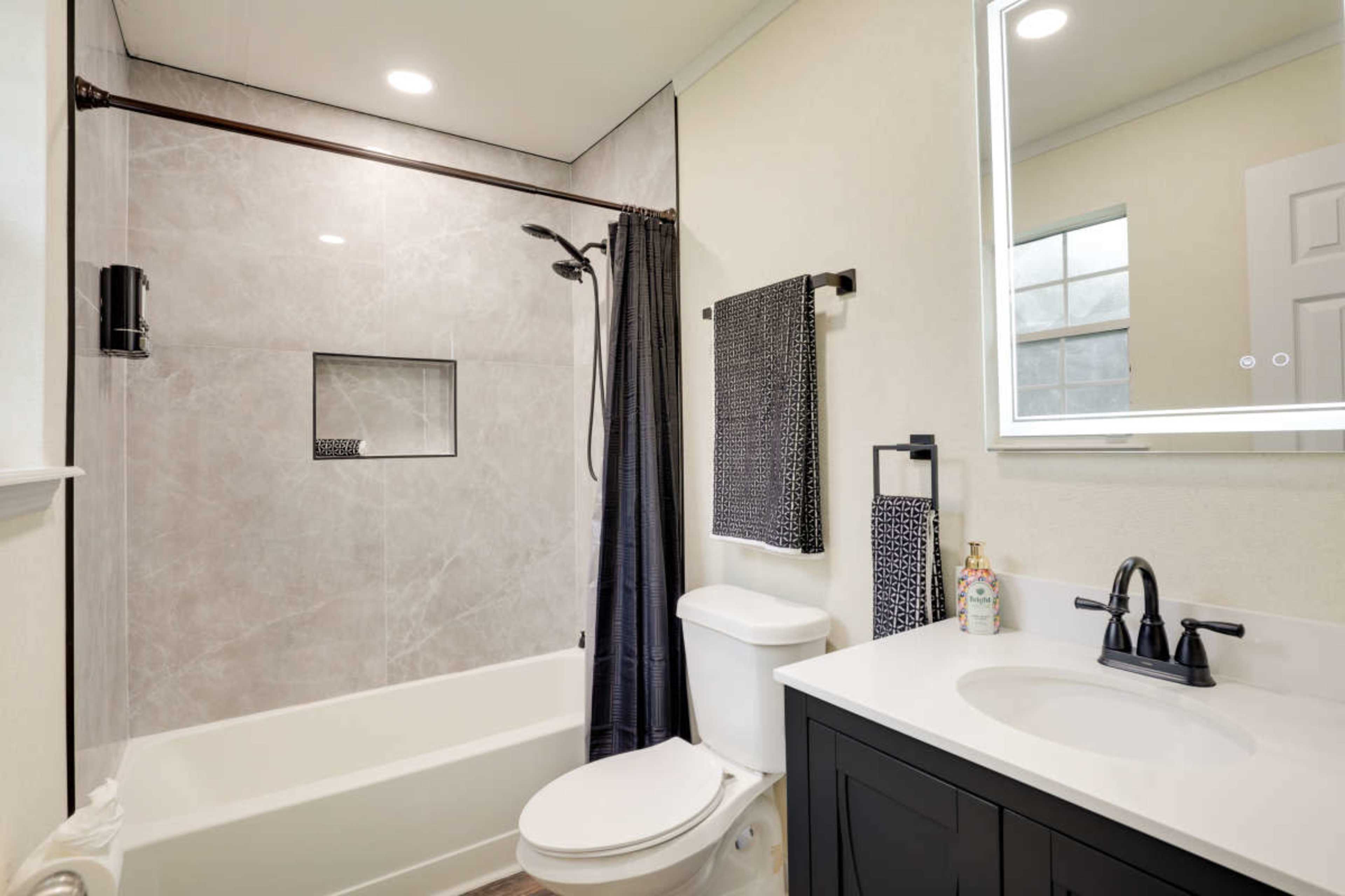 A modern bathroom featuring a shower-tub combination, a toilet, a black vanity with a white countertop, and a large mirror.