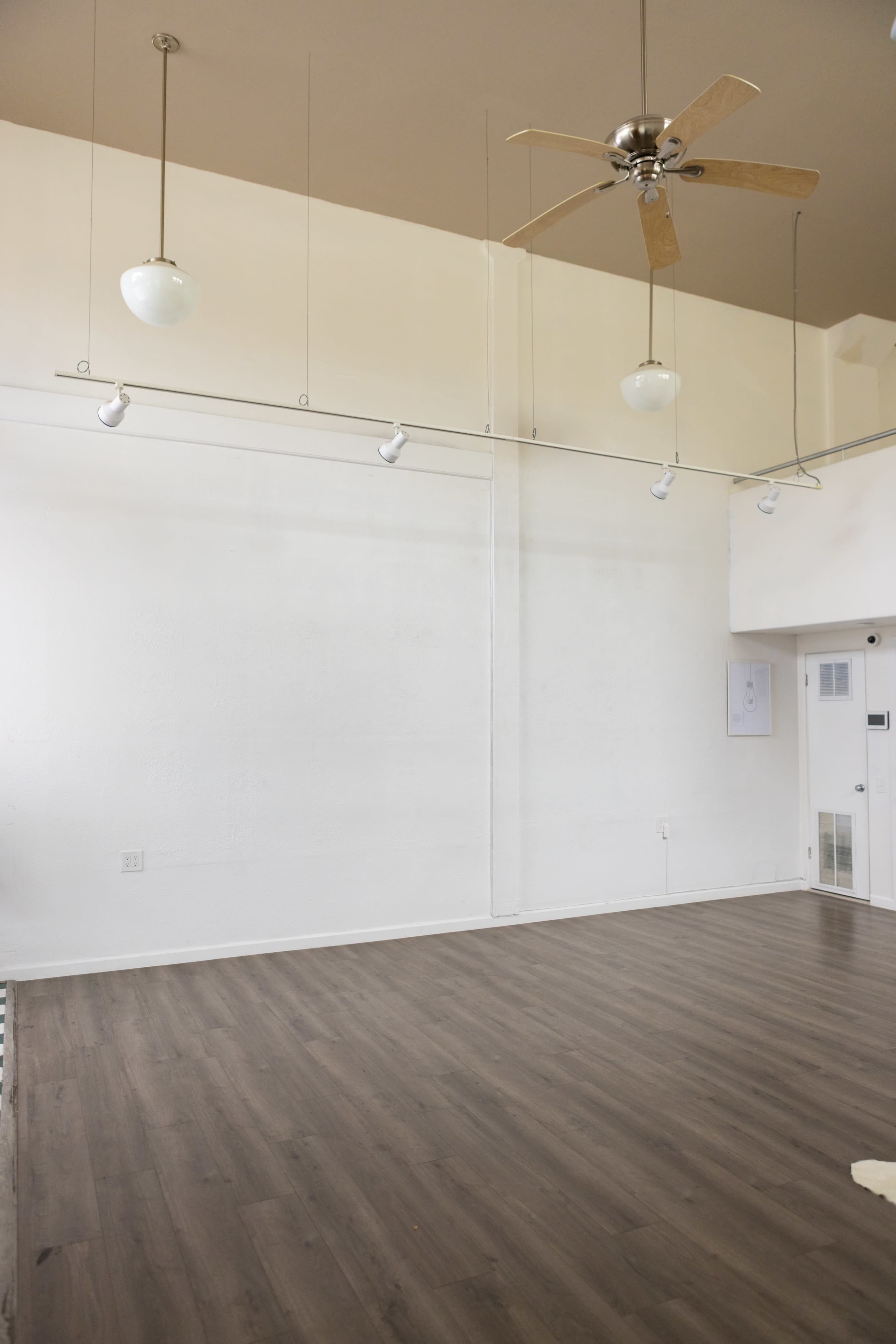 The image shows an empty room with a wooden floor, white walls, and a ceiling fan, illuminated by hanging light fixtures.