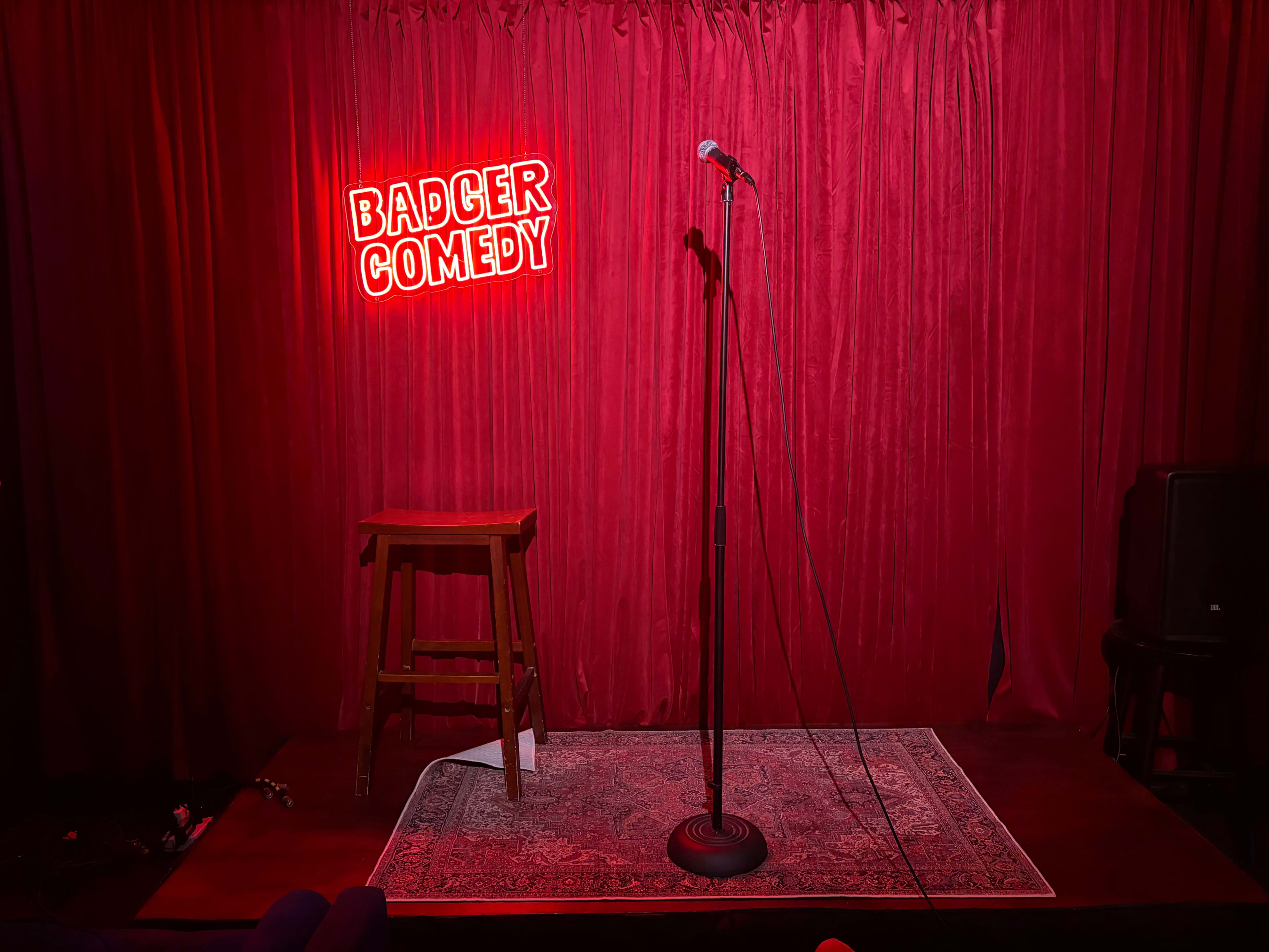 A microphone stands on a stage in front of a red velvet curtain, next to a wooden stool and a neon sign that reads "BADGER COMEDY."