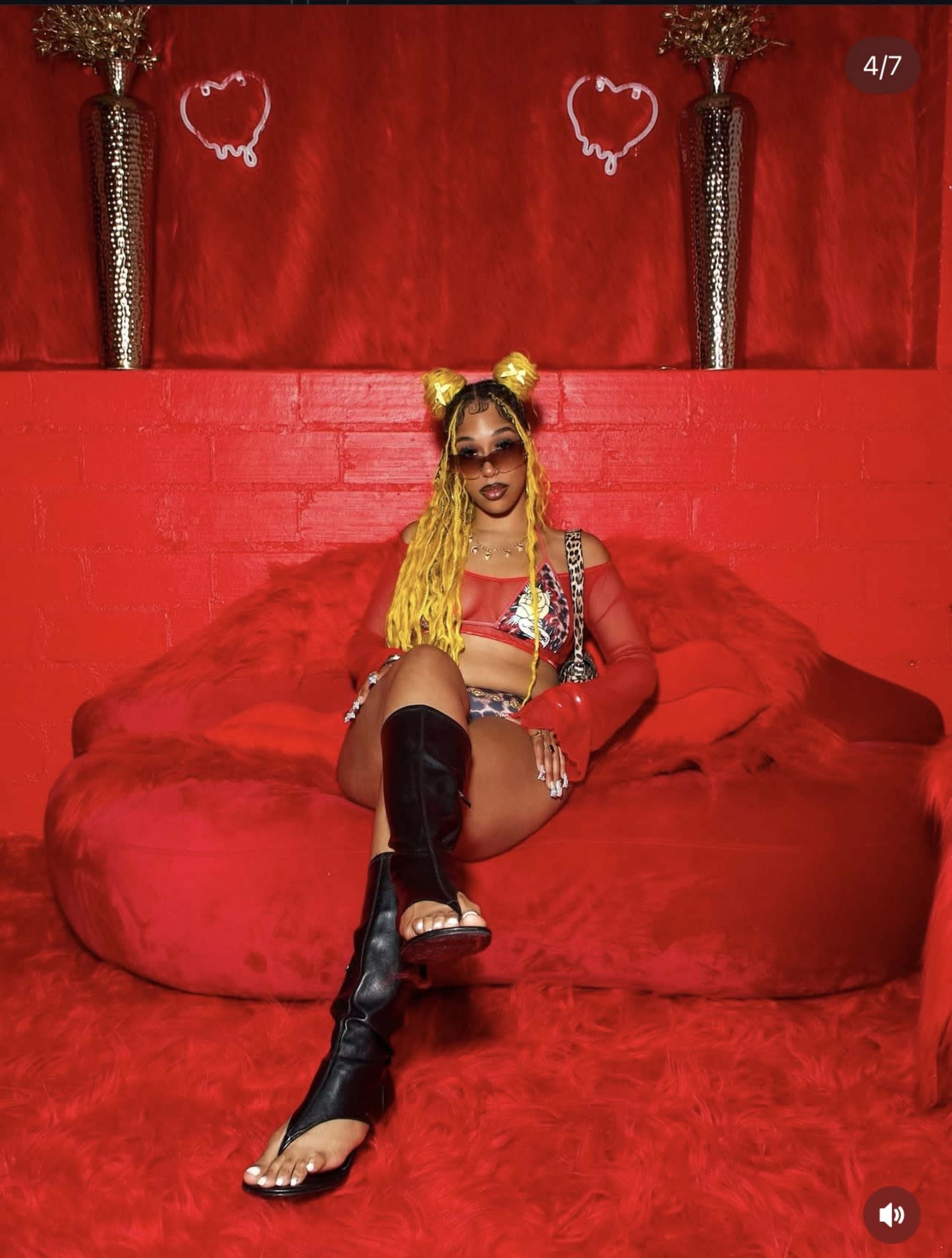 A person with long, yellow braided hair and sunglasses sits on a red couch against a red wall decorated with heart motifs.
