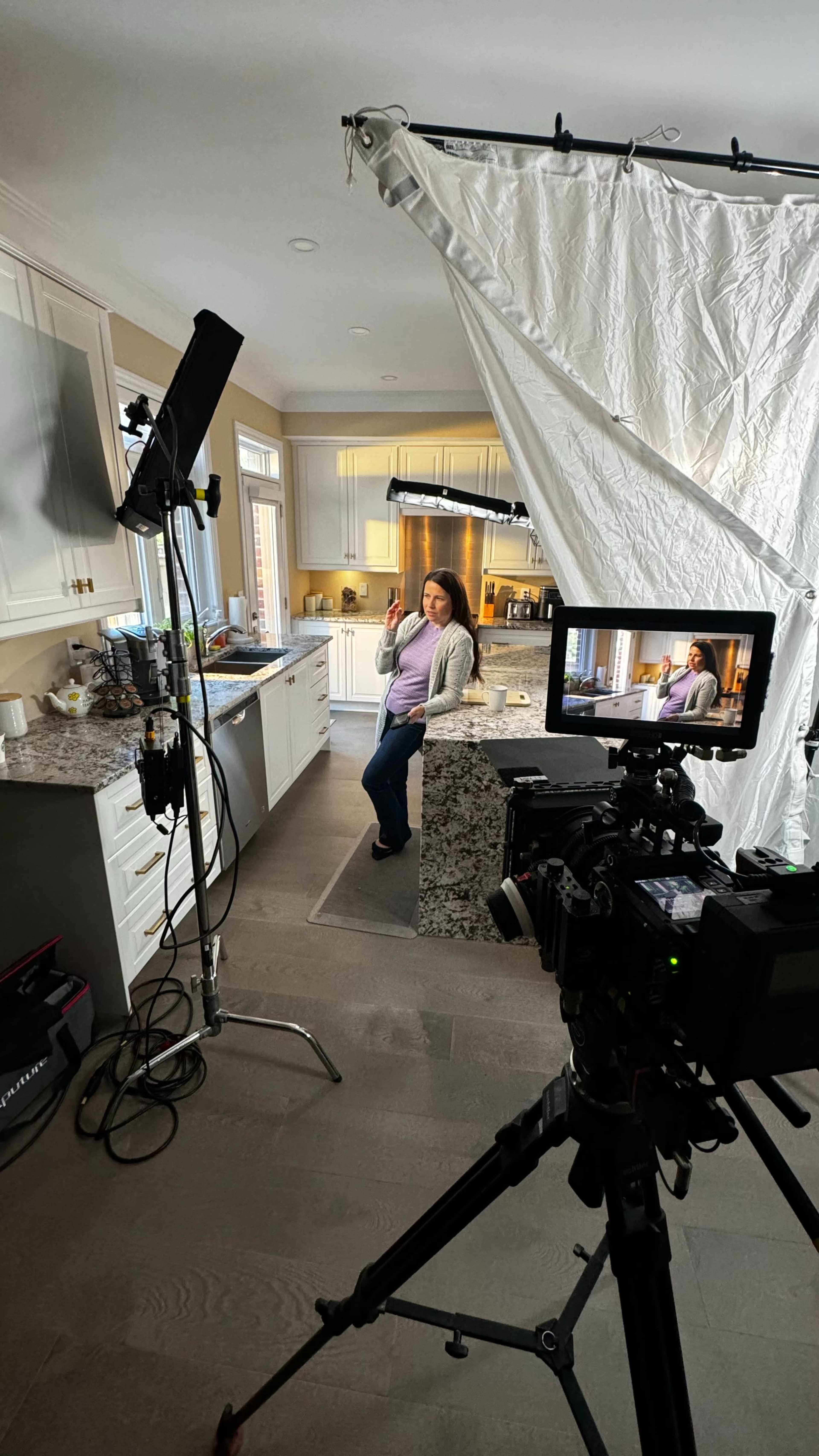 A woman stands in a kitchen while a camera and lighting equipment are set up for a shoot.