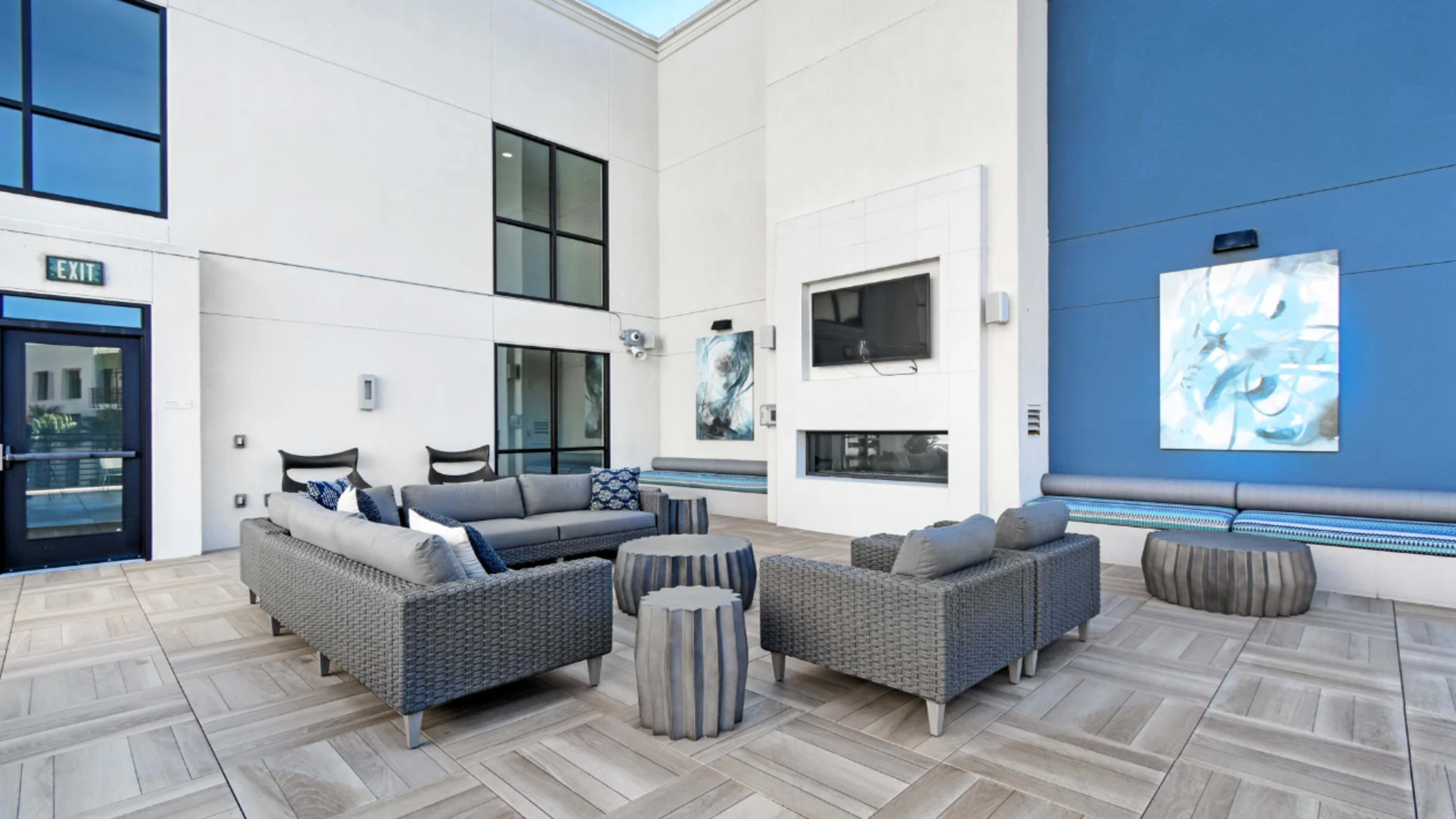A modern lounge area features a set of gray wicker seating around a central table, with large windows and blue accent walls.