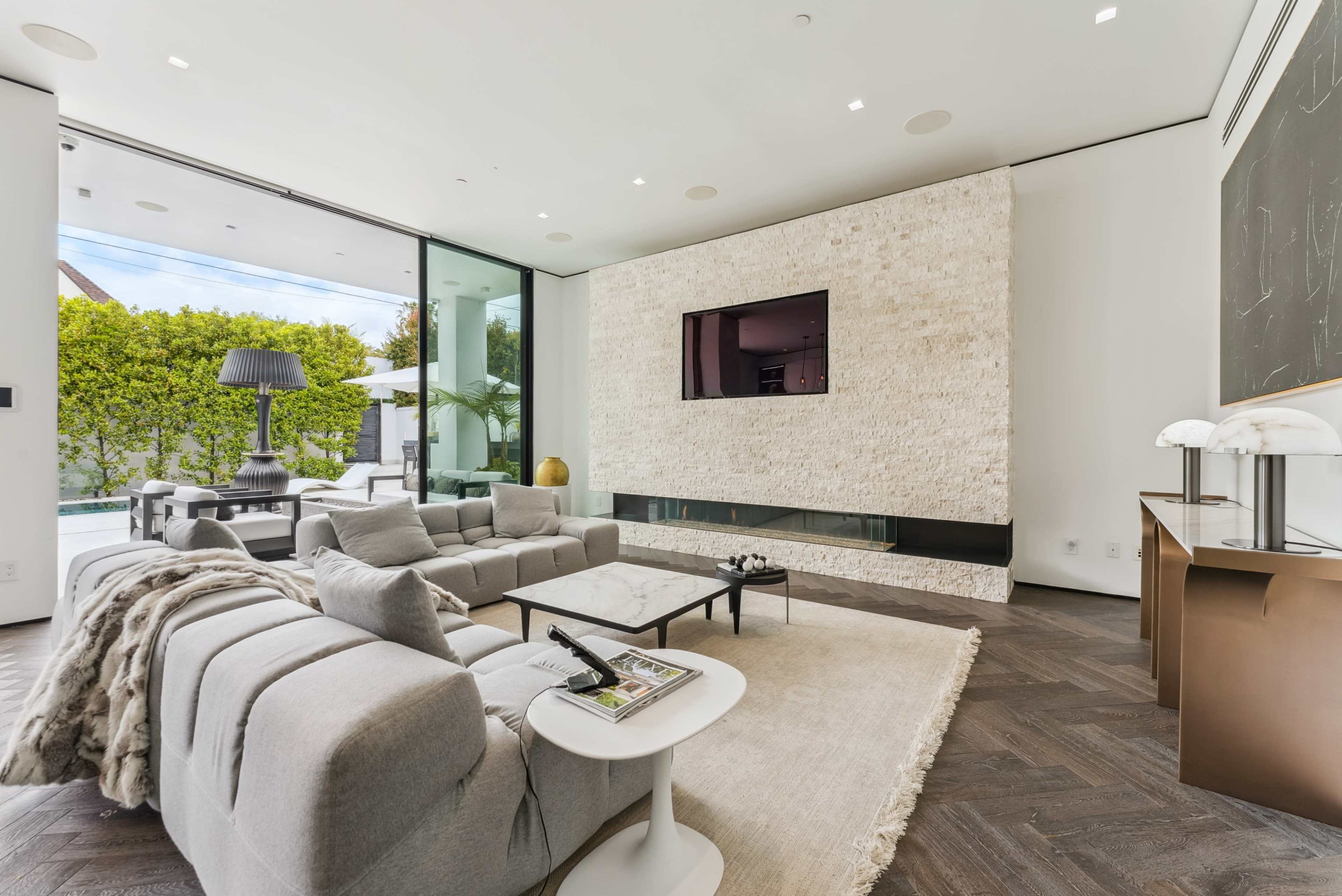 The modern living room features a light-colored sofa set, a flat-screen TV mounted on a textured wall, and large glass doors opening to an outdoor area.