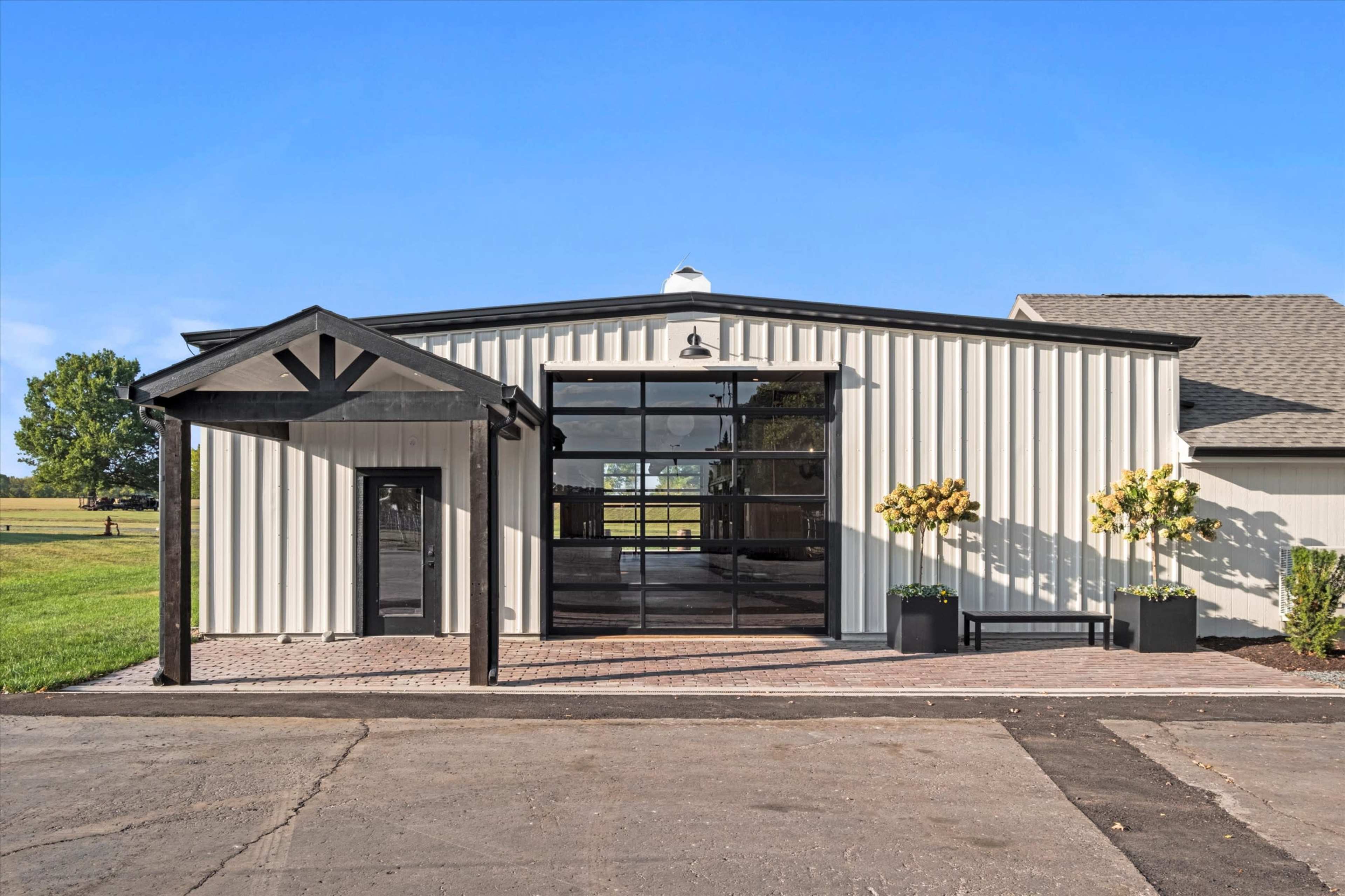 The image shows a modern, metal-clad building with large glass panels and a covered entrance, surrounded by greenery and a paved area.