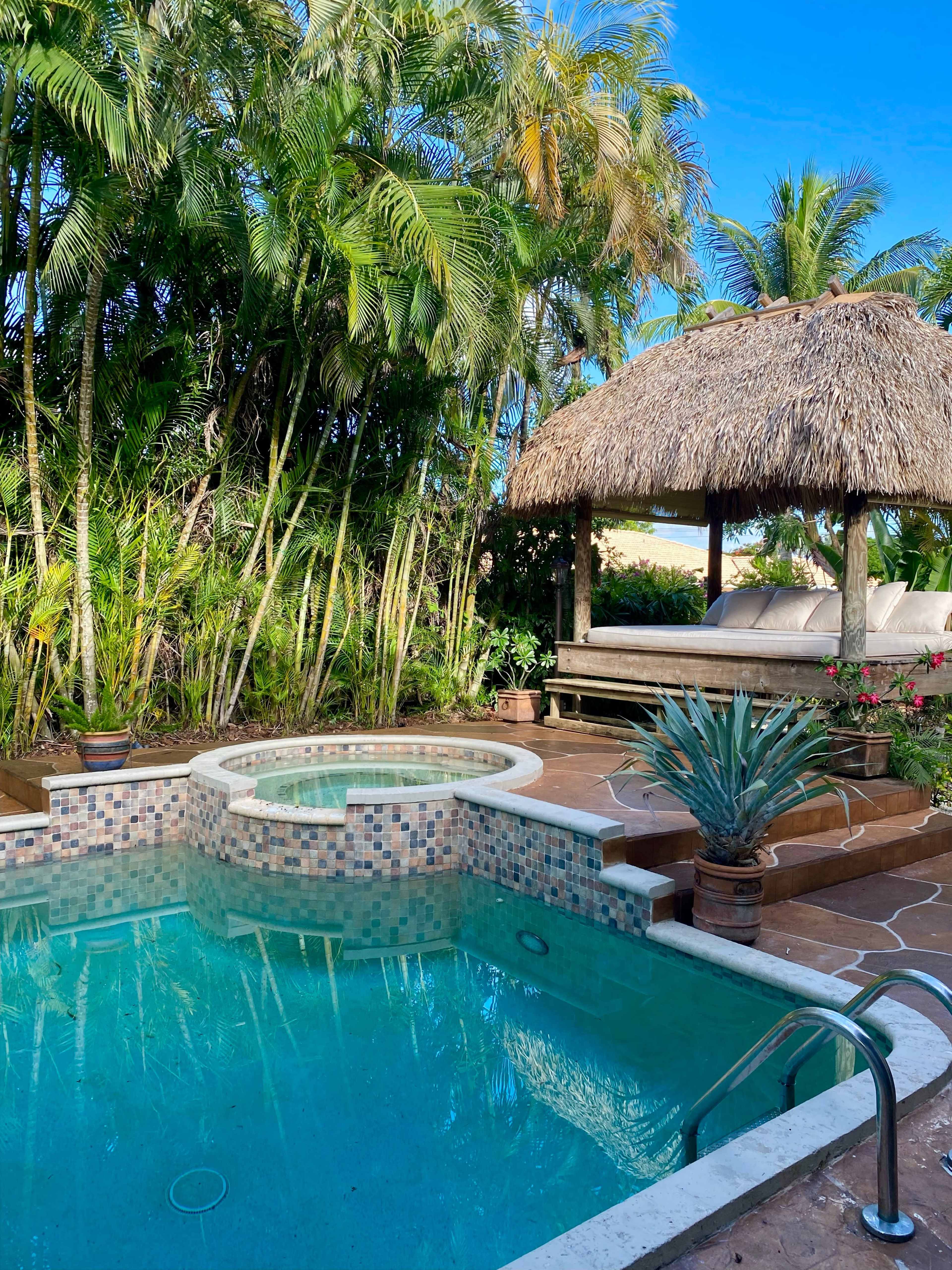 A sparkling hot tub is surrounded by lush palm trees and a thatched-roof gazebo with a couch.