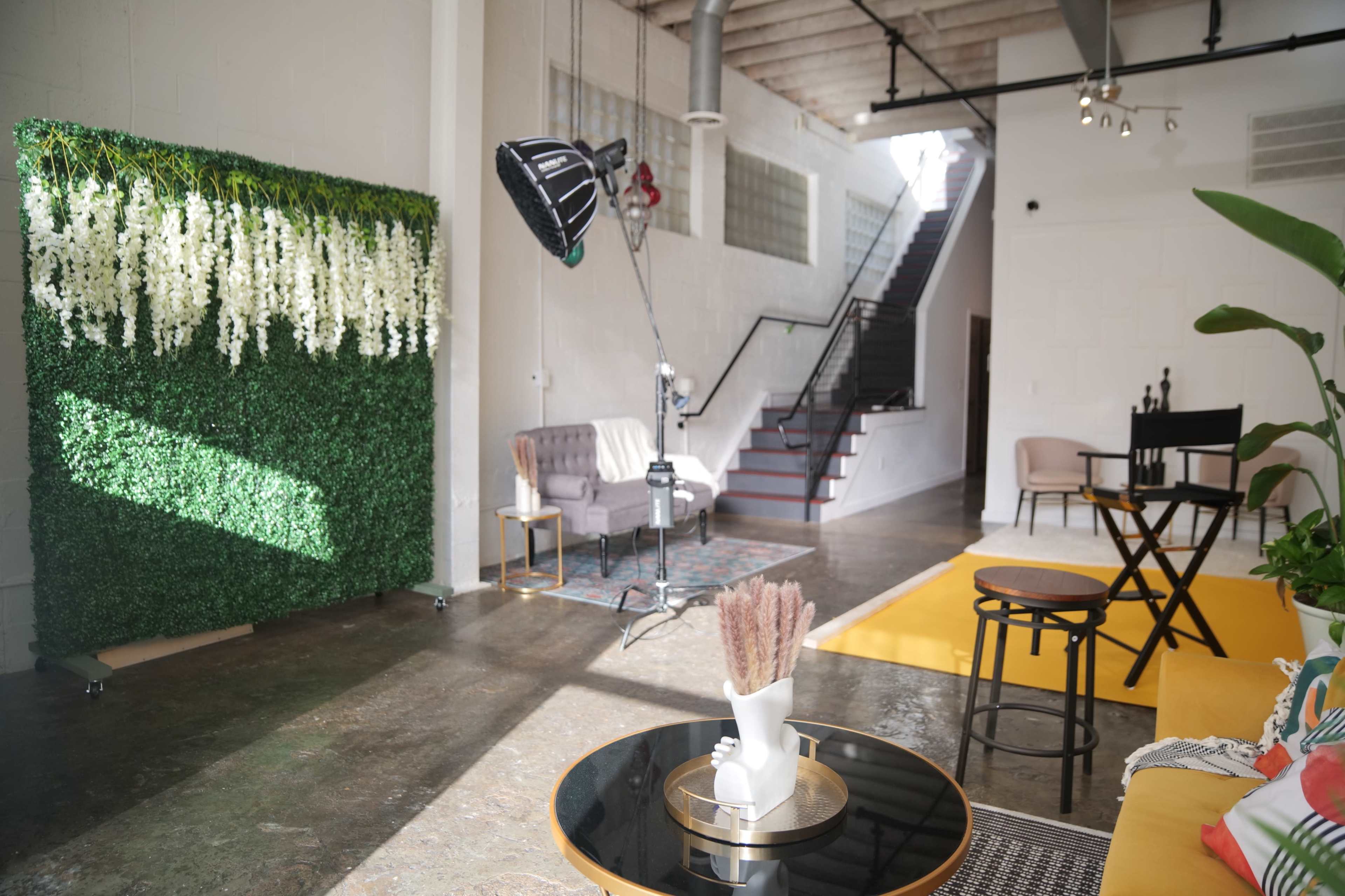 A modern studio space featuring a green wall of artificial greenery, a seating area with a sofa, a small table, and a large window allowing natural light to enter.