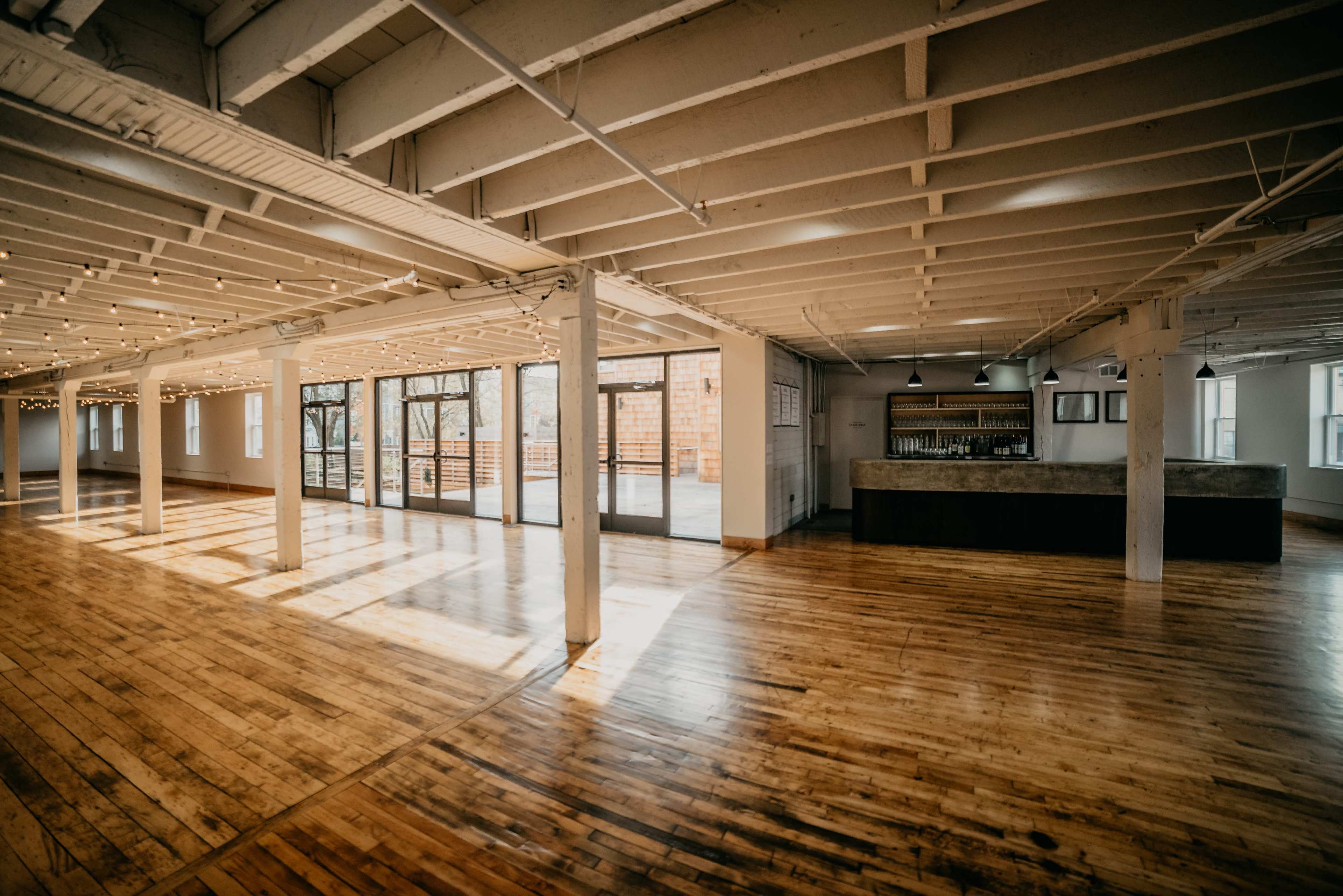 The image shows a spacious, well-lit interior space with wooden flooring and large windows, featuring a bar area in one corner.