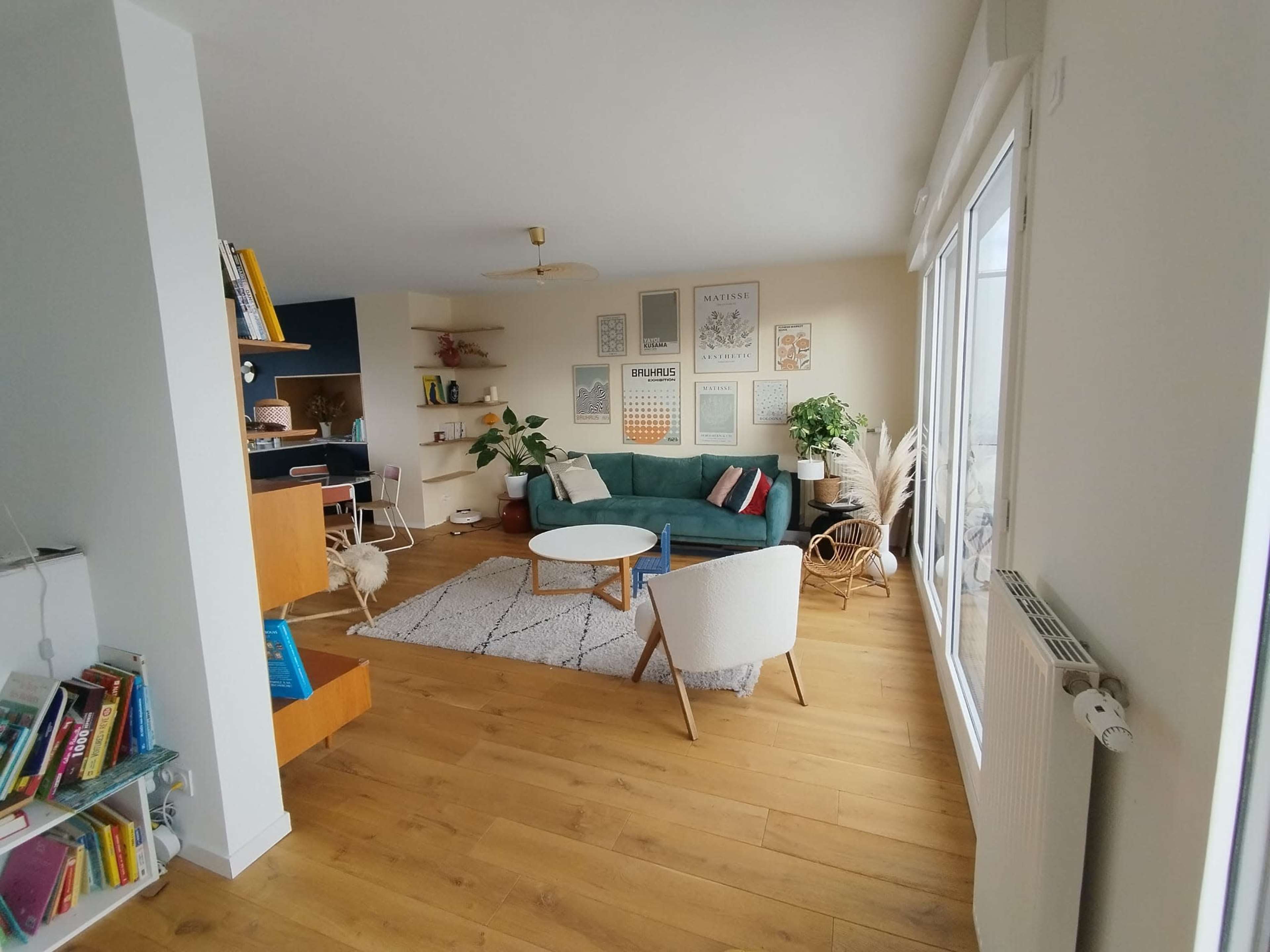A modern living room with a green sofa, a round coffee table, and a bookshelf, featuring wooden flooring and large windows.