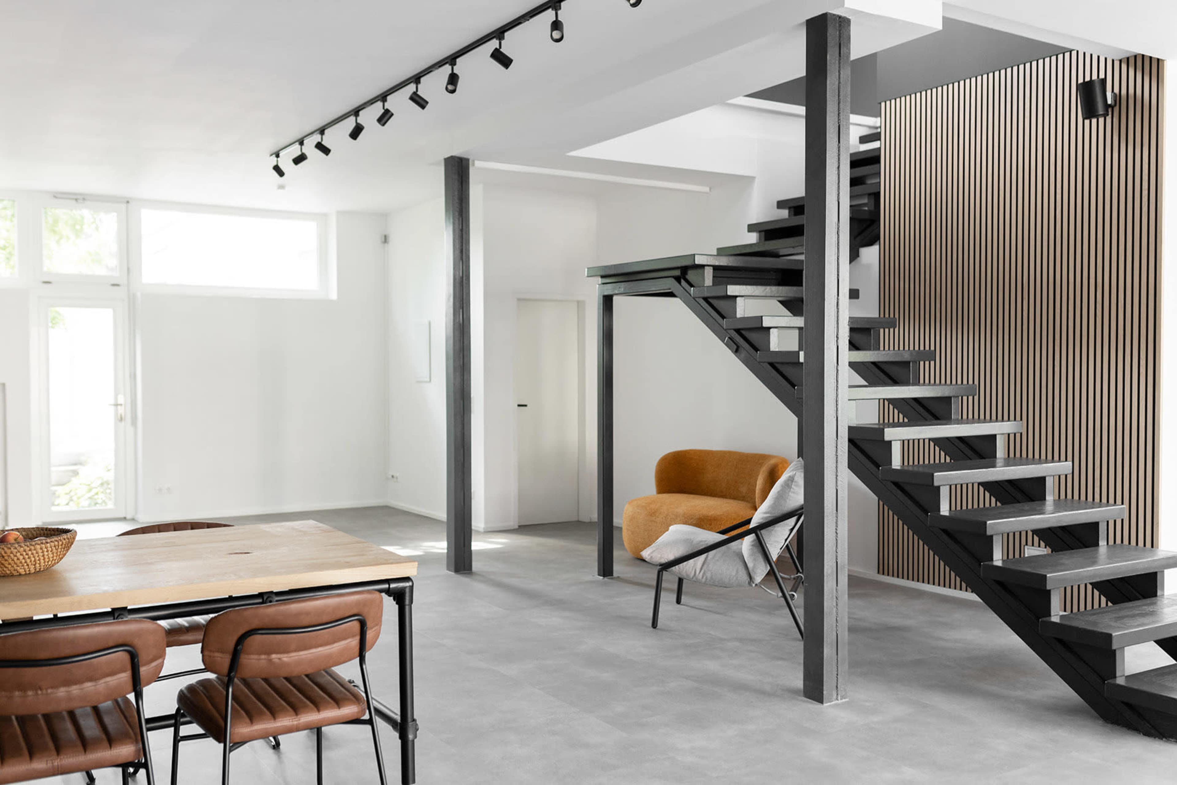 The image shows a contemporary interior space featuring a dining table with chairs, a staircase with a black metal frame, and large windows allowing natural light.