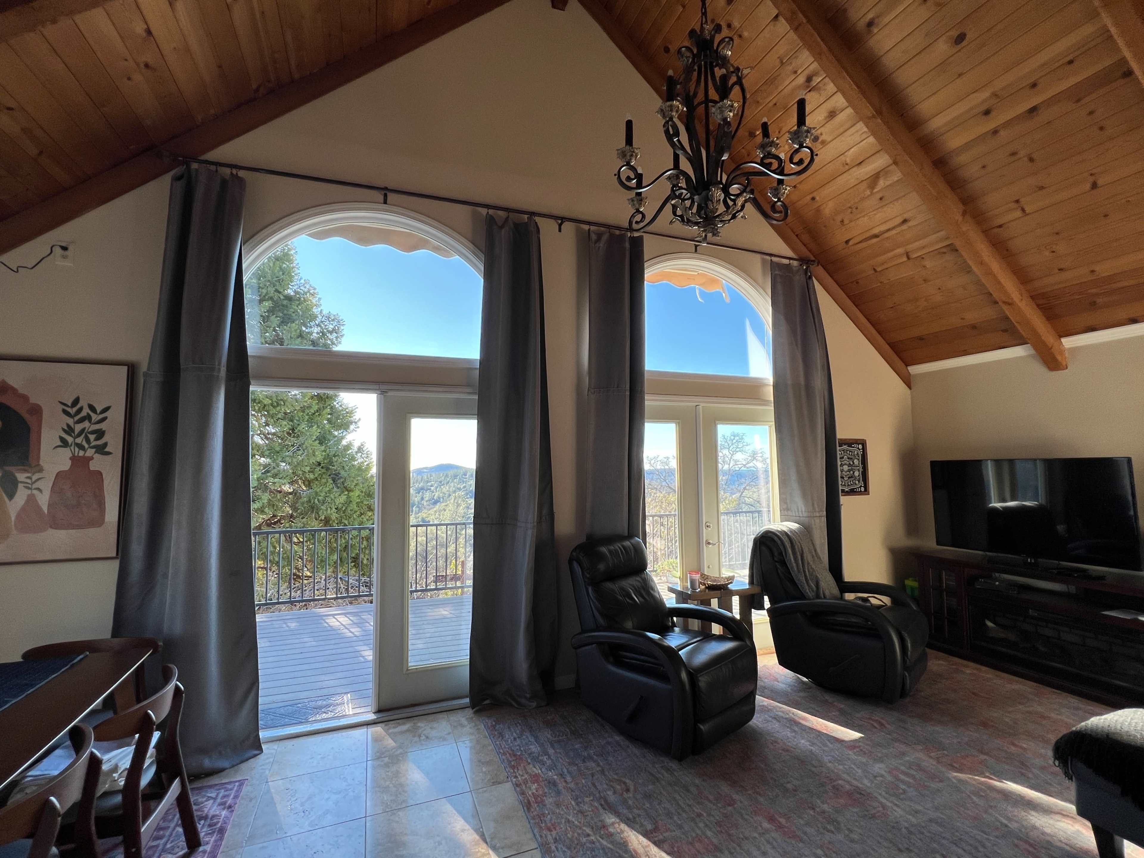 A spacious living room features two large arched windows with sheer curtains, two black recliners, and a TV against a wooden wall.