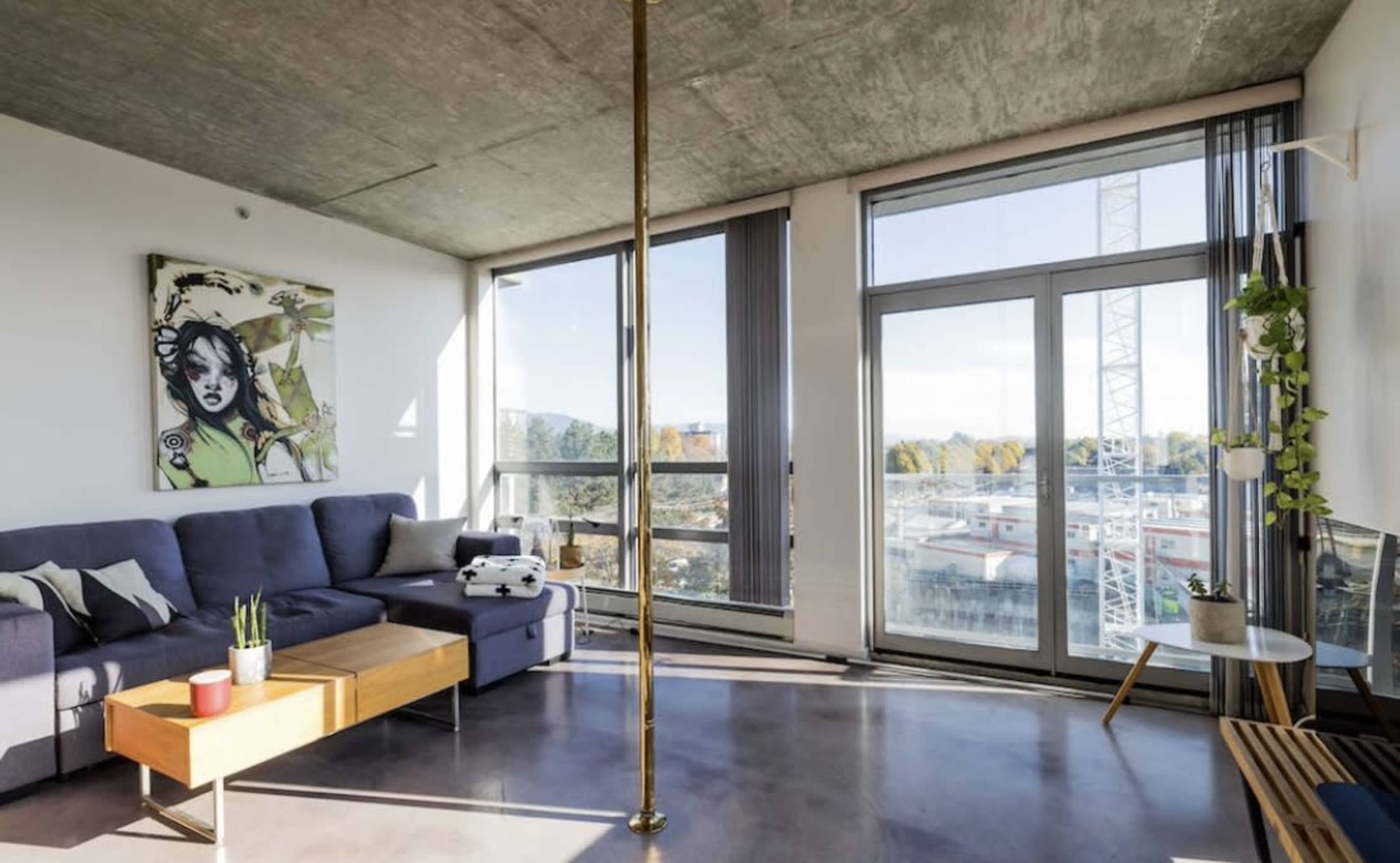 A modern living room featuring a sectional sofa, a wooden coffee table, a floor-to-ceiling window with a view, and a decorative pole.