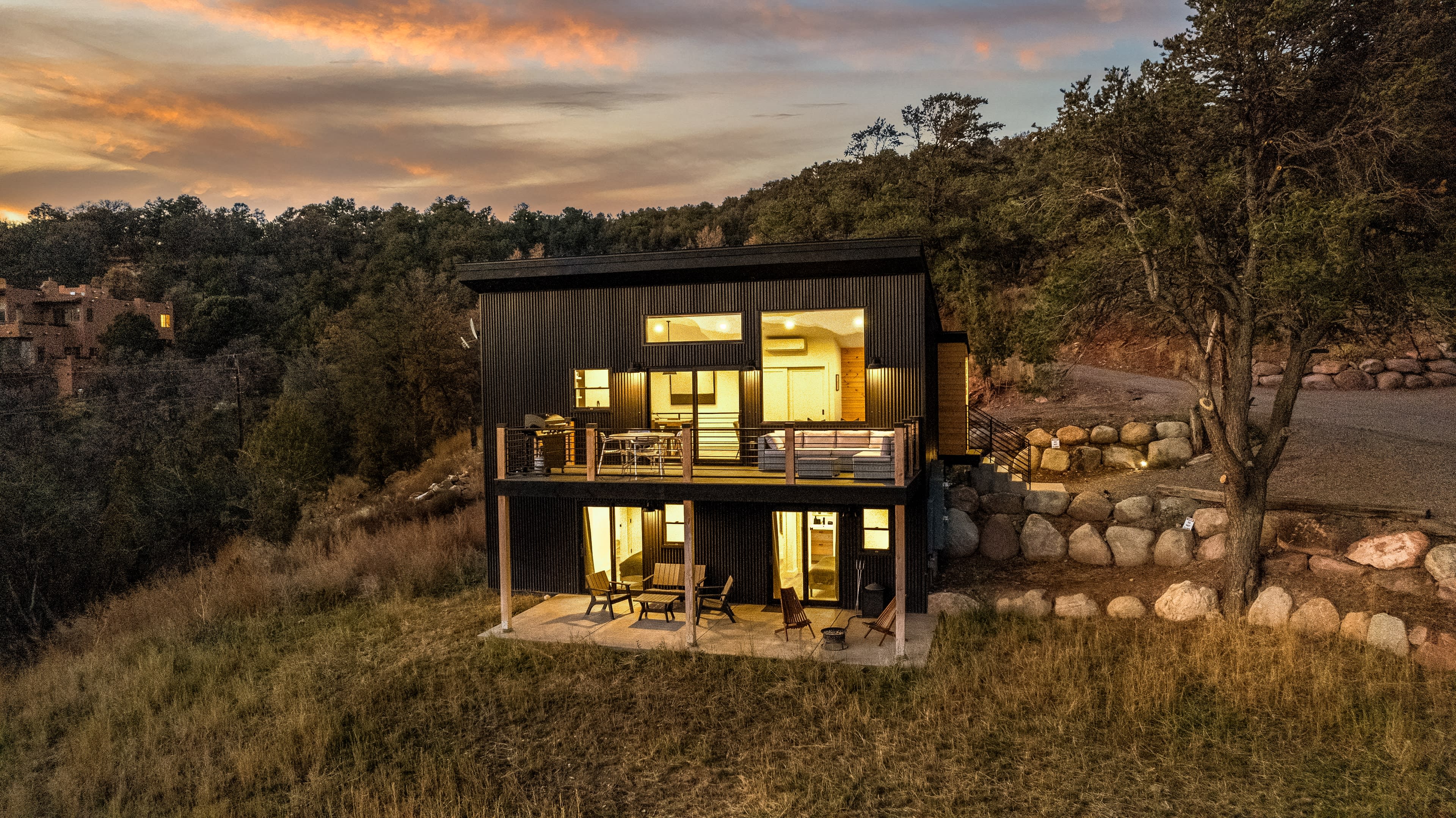 A modern two-story black house sits on a hillside, surrounded by grass and trees, with a balcony overlooking the landscape at sunset.