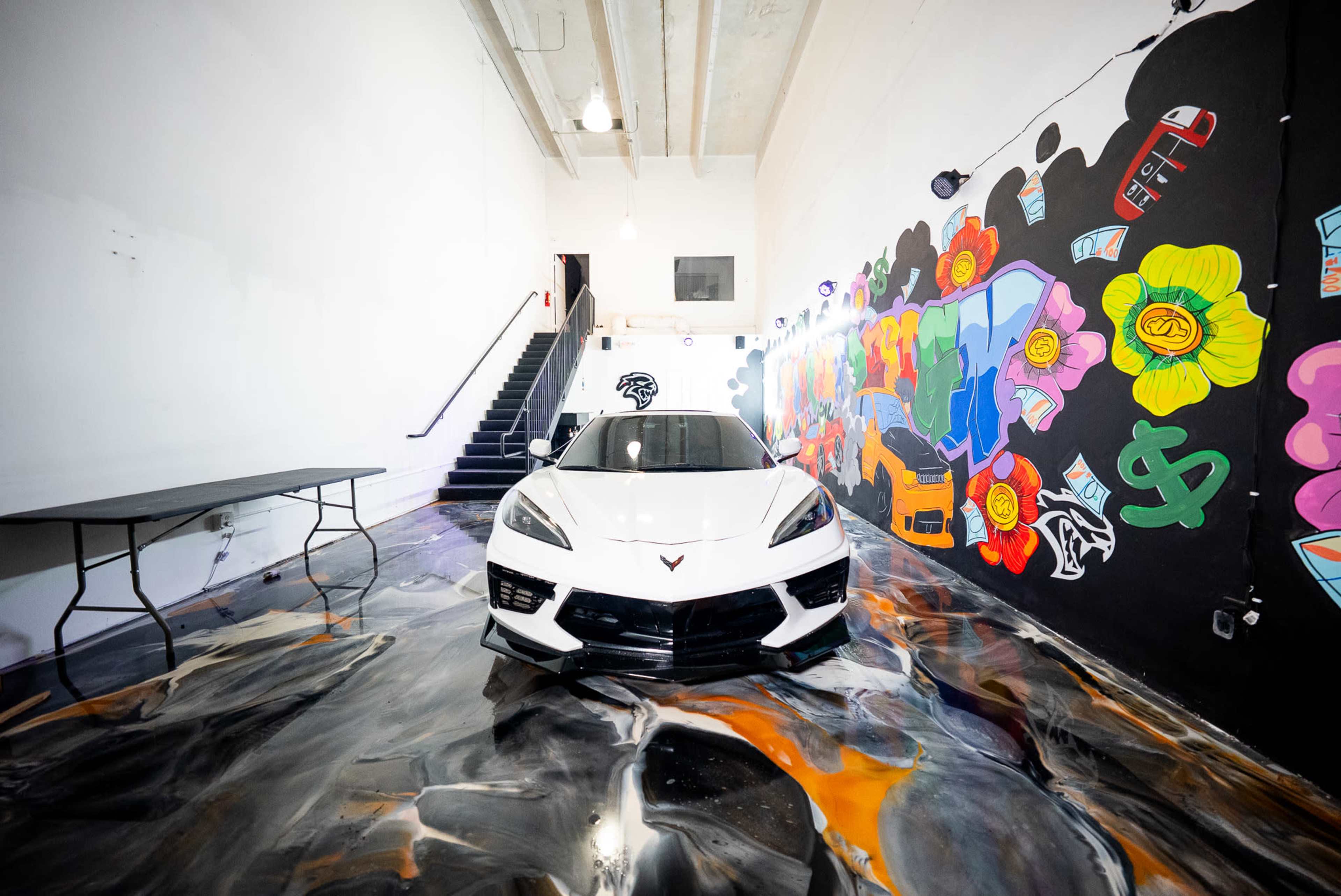 A white sports car is parked in a colorful room with a mural on one wall and a staircase in the background.