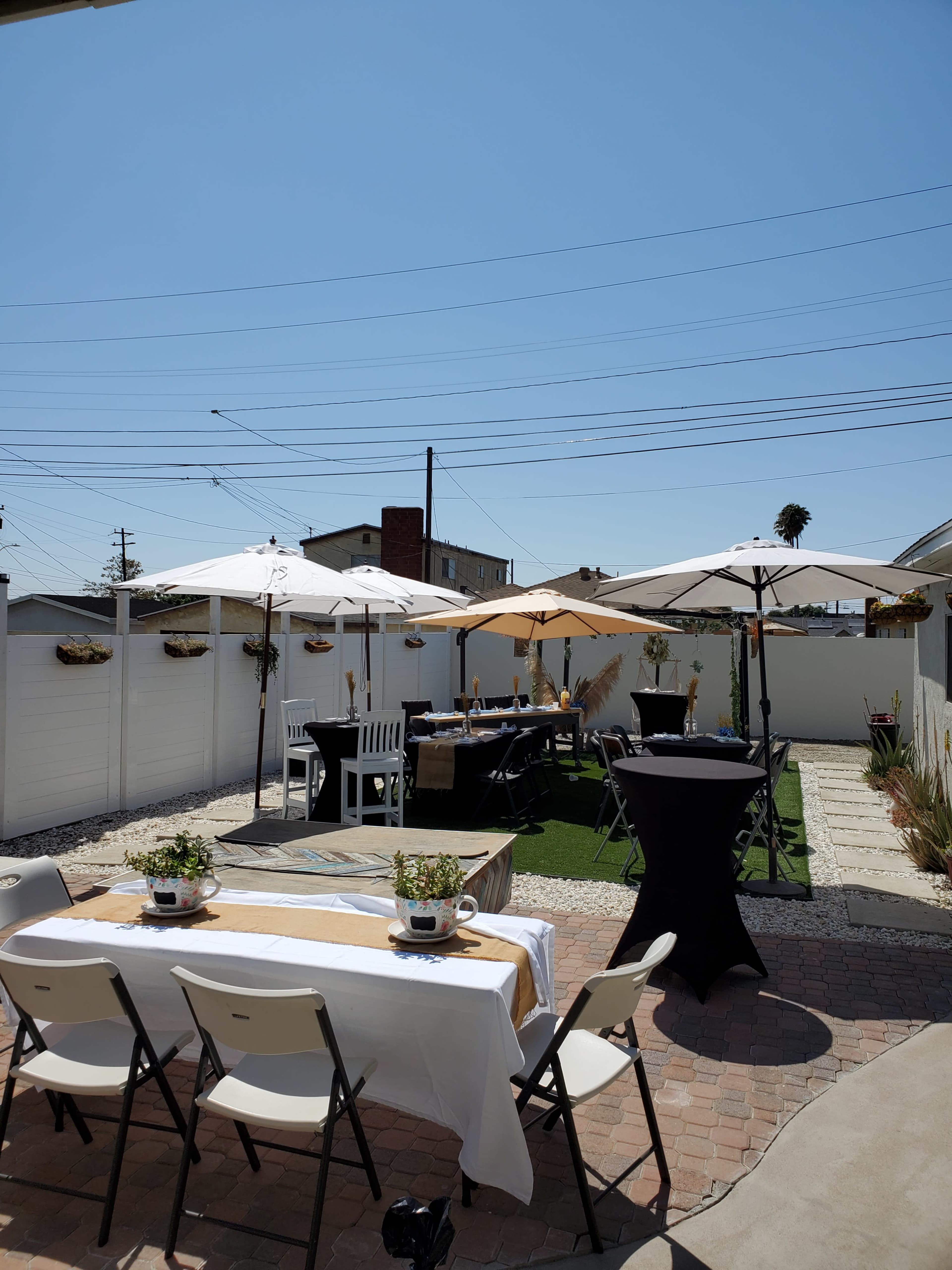 A sunlit outdoor patio features several tables with umbrellas, decorated with plants and surrounded by a white fence.
