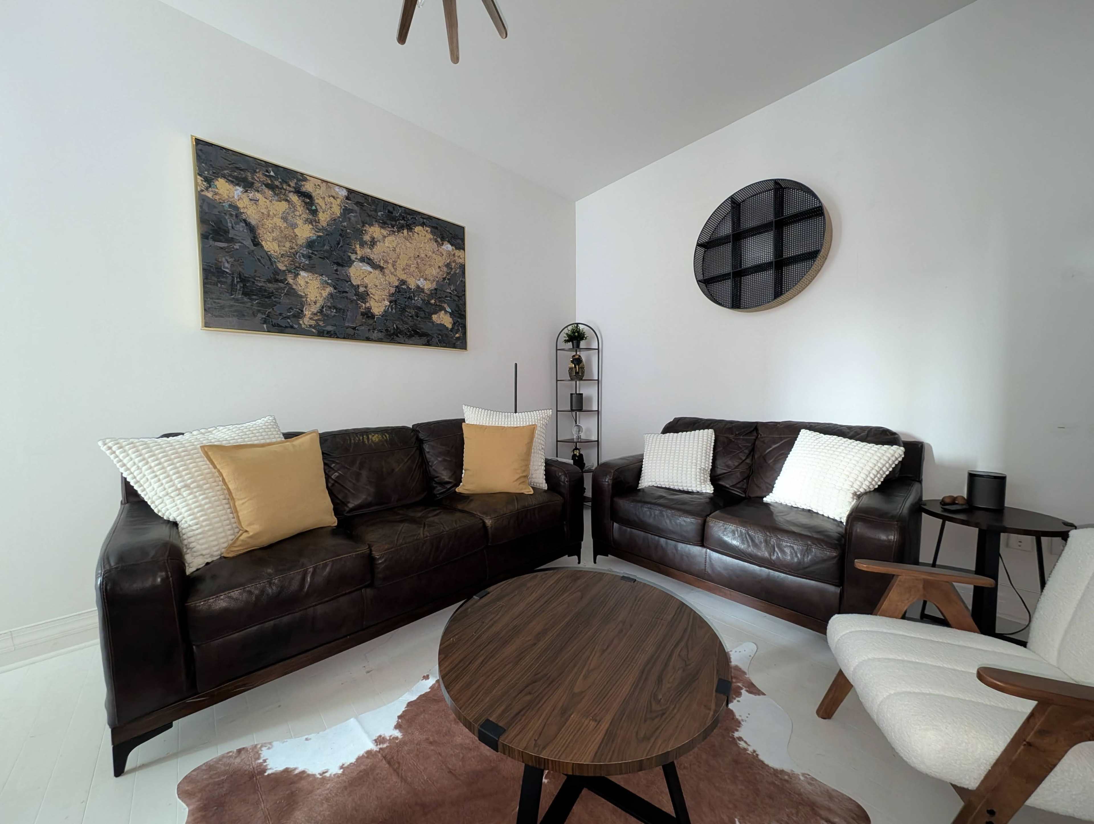 The image shows a modern living room with two dark leather sofas, a wooden coffee table, a geometric wall art piece, and a small bookshelf.