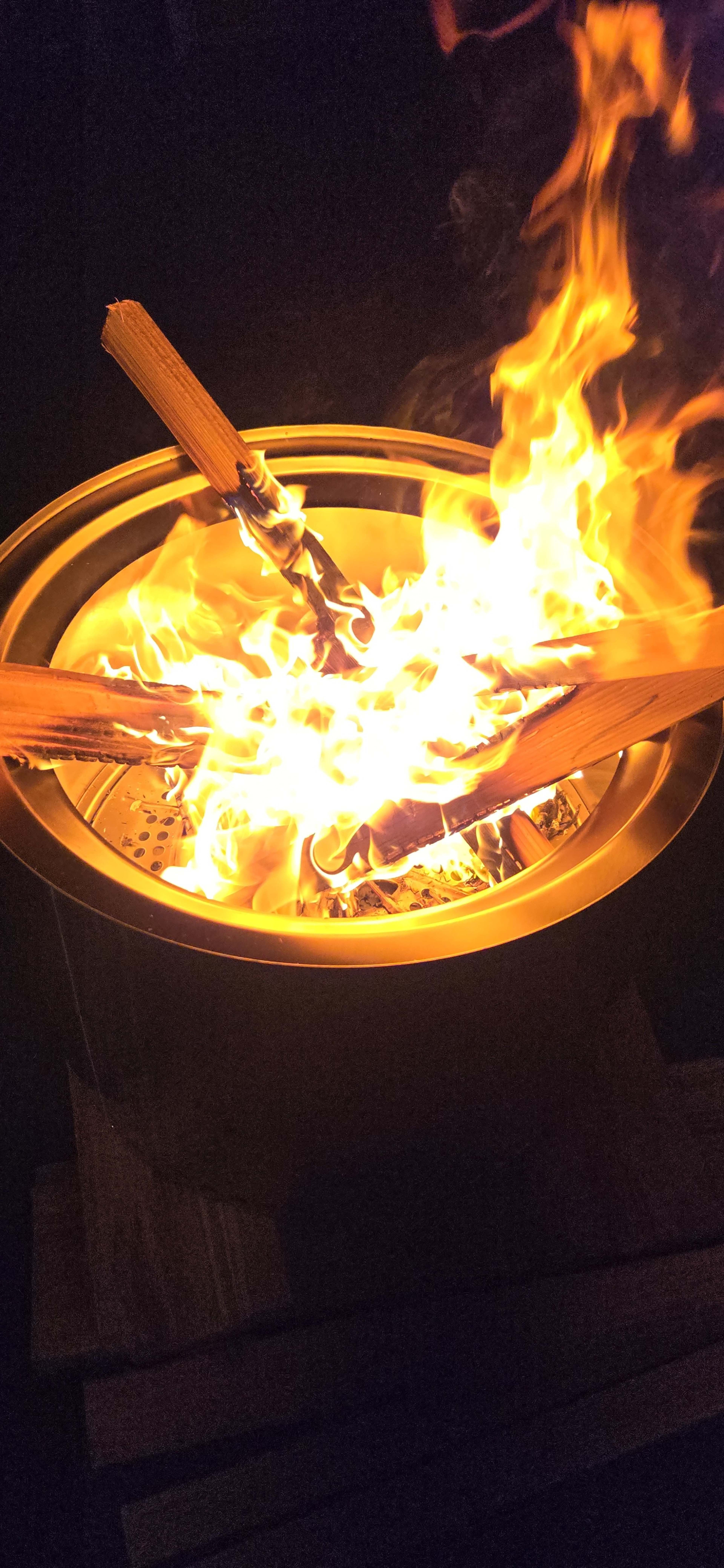 A fire burns brightly in a circular metal fire pit, with wooden logs crackling and glowing amid the flames.