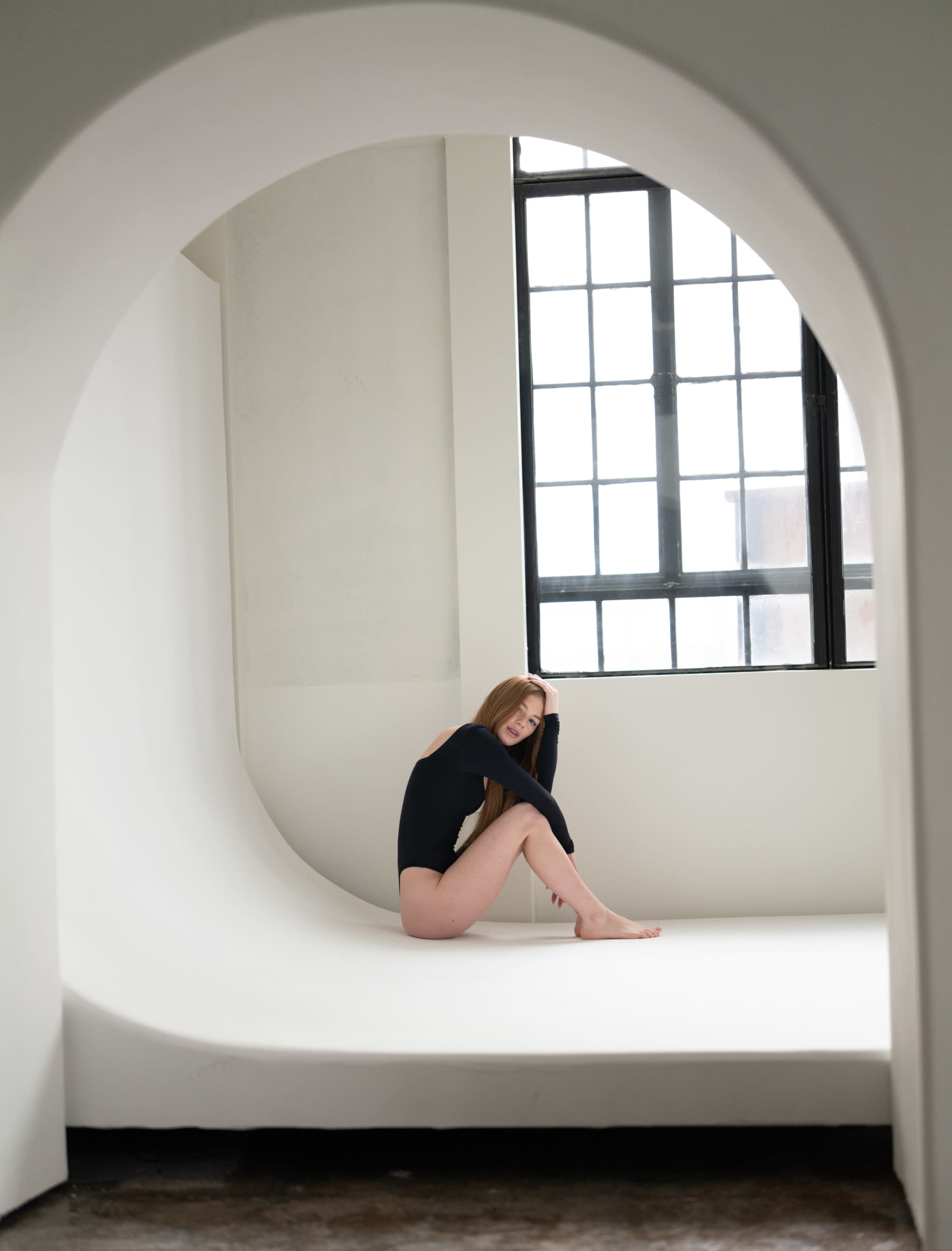 A person sits in a minimalist, curved white space near a large window with black frames.