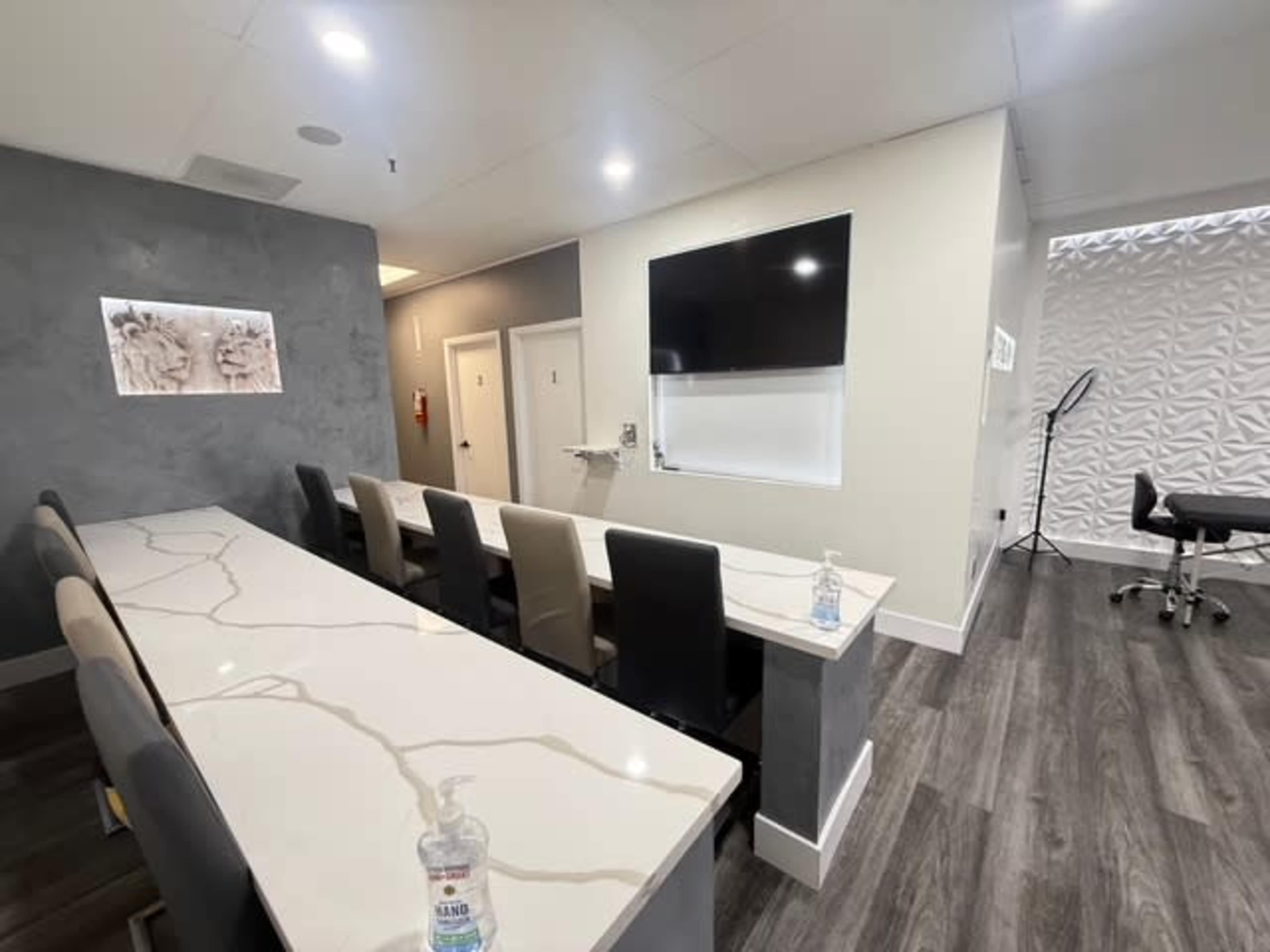 The image shows a modern, well-lit interior space featuring a long, marble-topped table with multiple chairs, a wall-mounted television, and a textured panel wall in the background.
