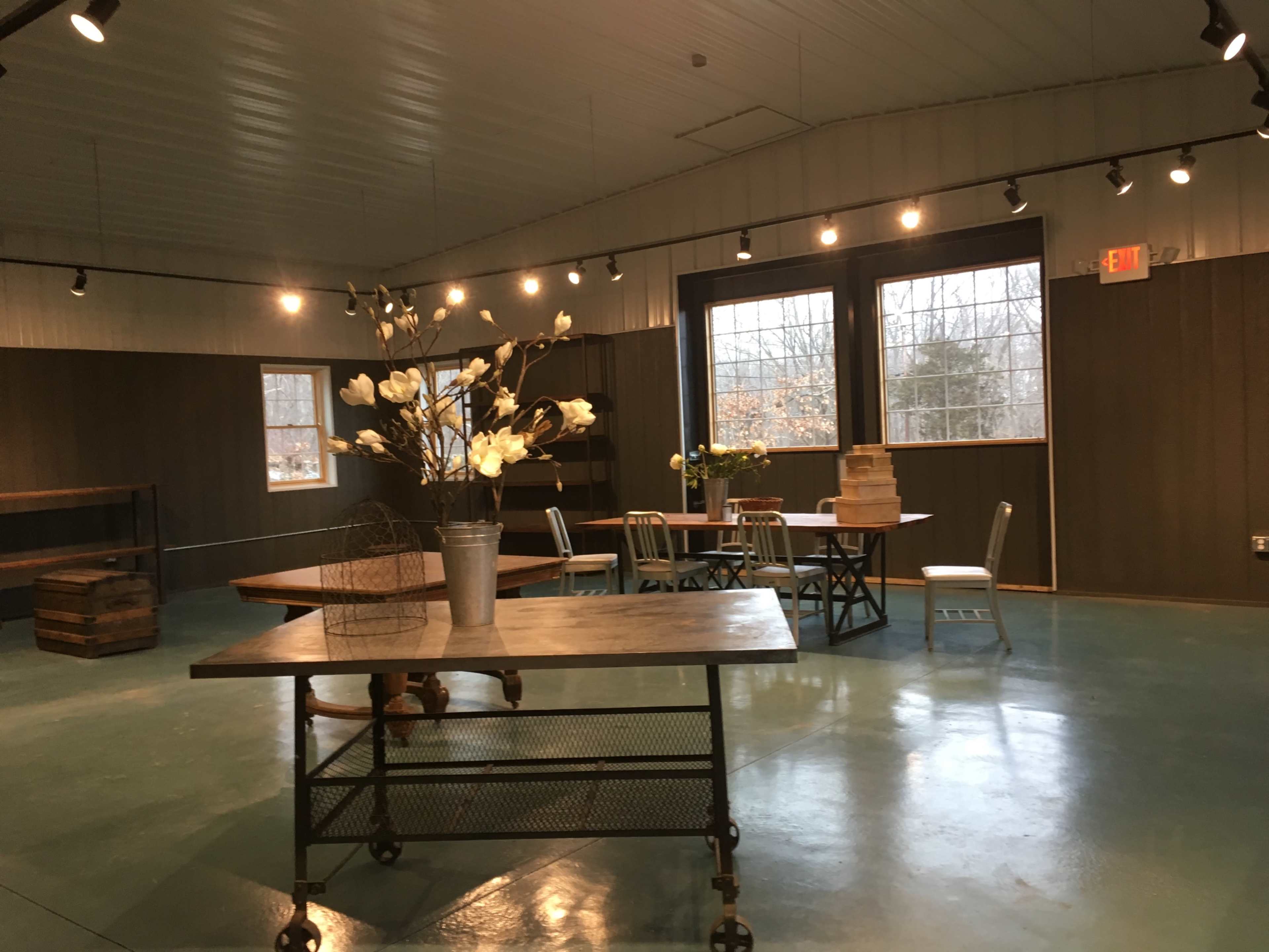 A spacious room with wooden tables, chairs, and decorative flowers, illuminated by overhead lighting and large windows.