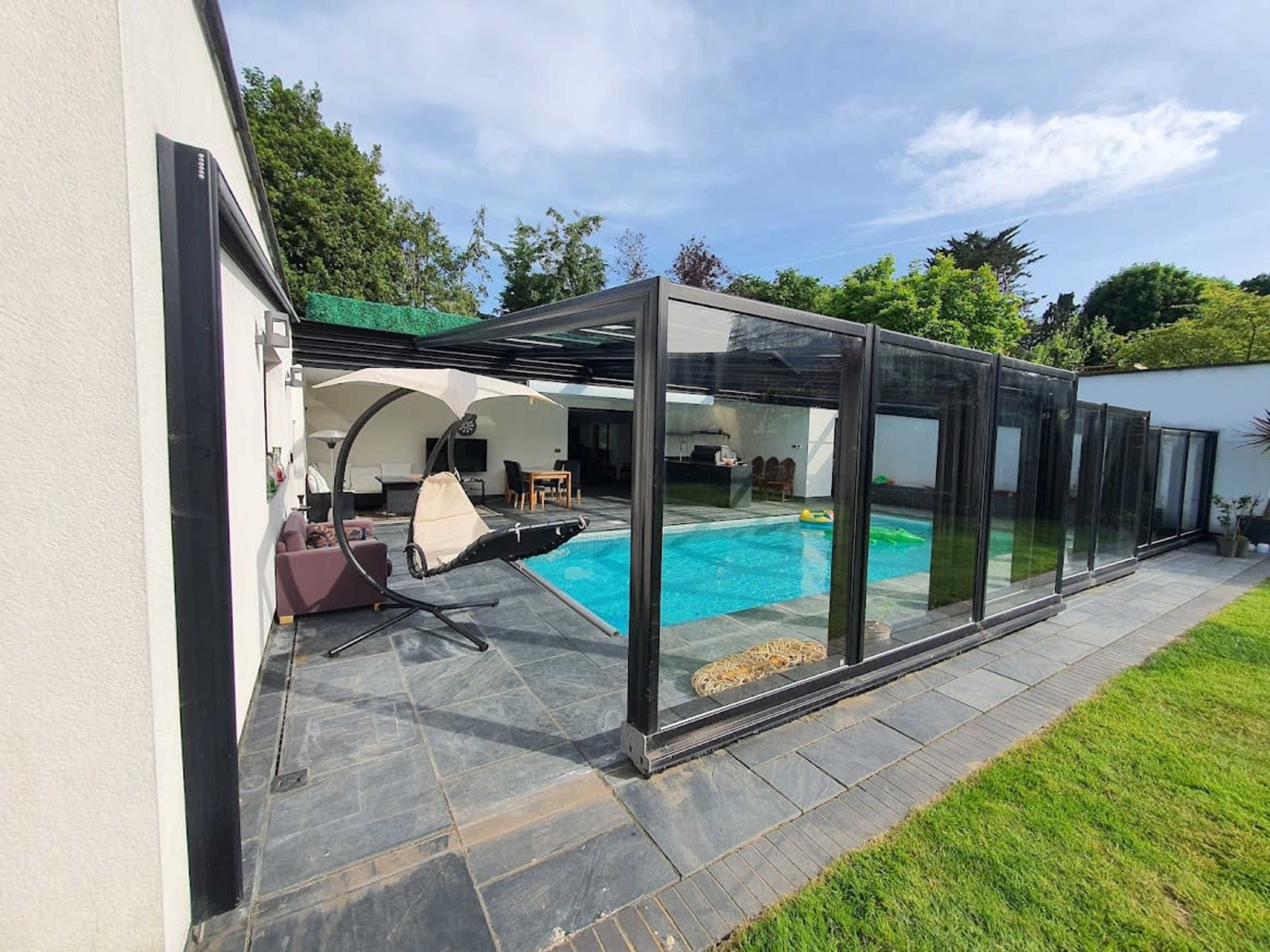 The image shows a modern pool area enclosed with glass panels, featuring a swimming pool surrounded by stone tiles and a grassy lawn.