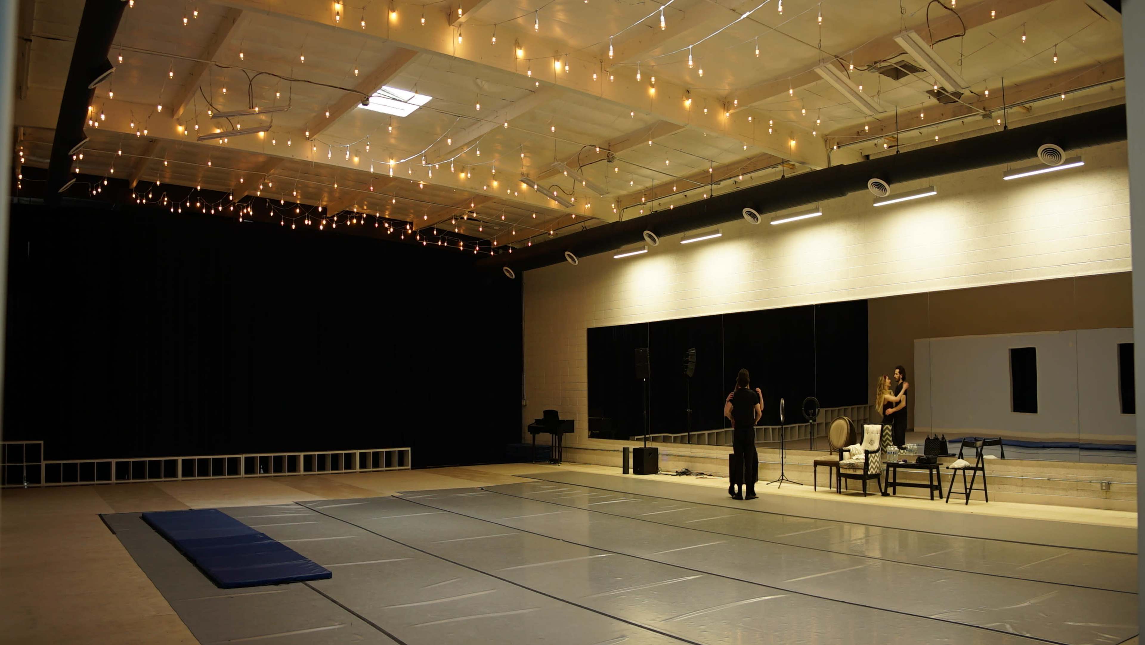 A spacious dance studio features a mirrored wall, a large clear floor area, and warm, ambient lighting from string lights hanging overhead.
