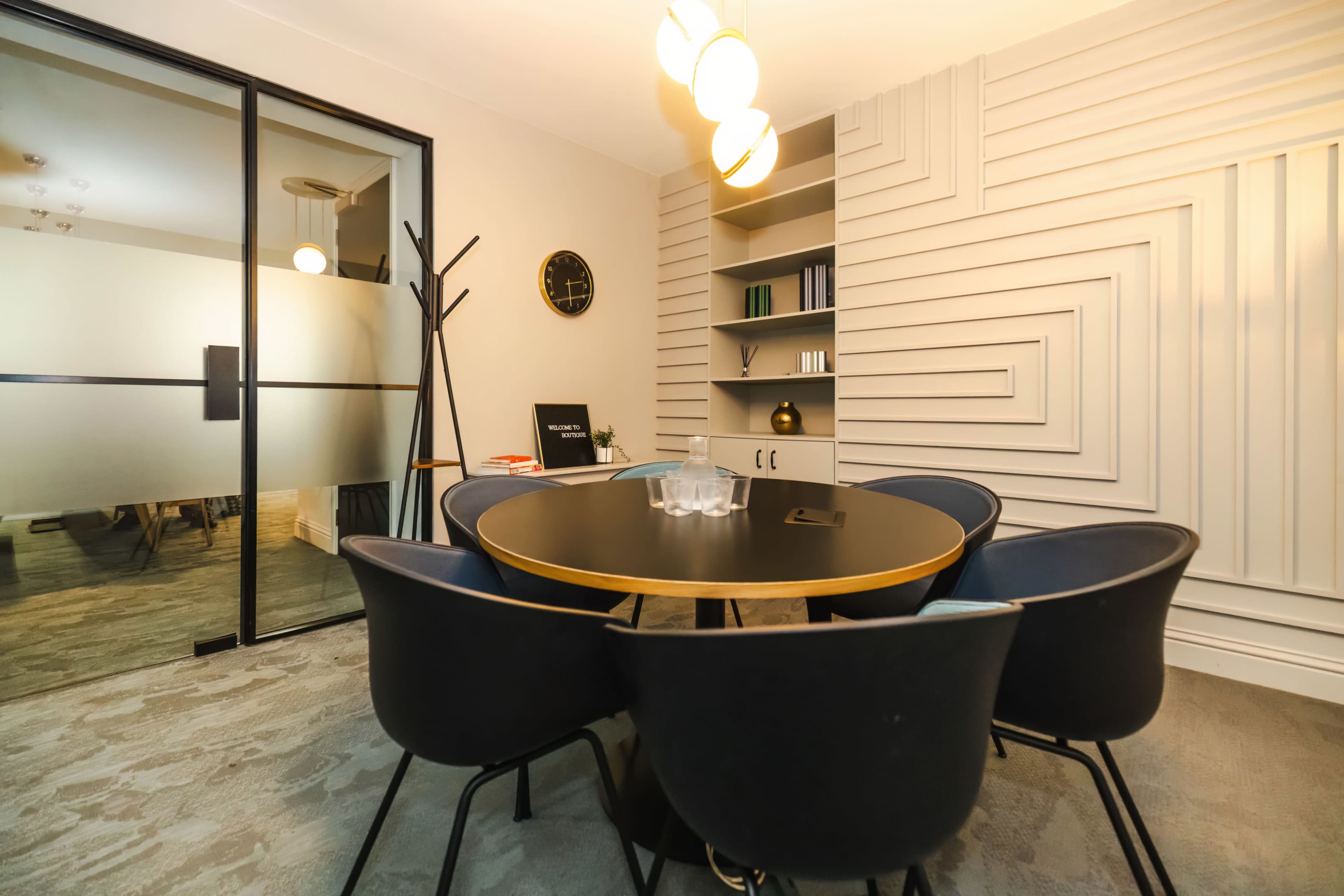 A modern conference room features a round black table surrounded by four blue chairs, with a glass partition and shelves in the background.