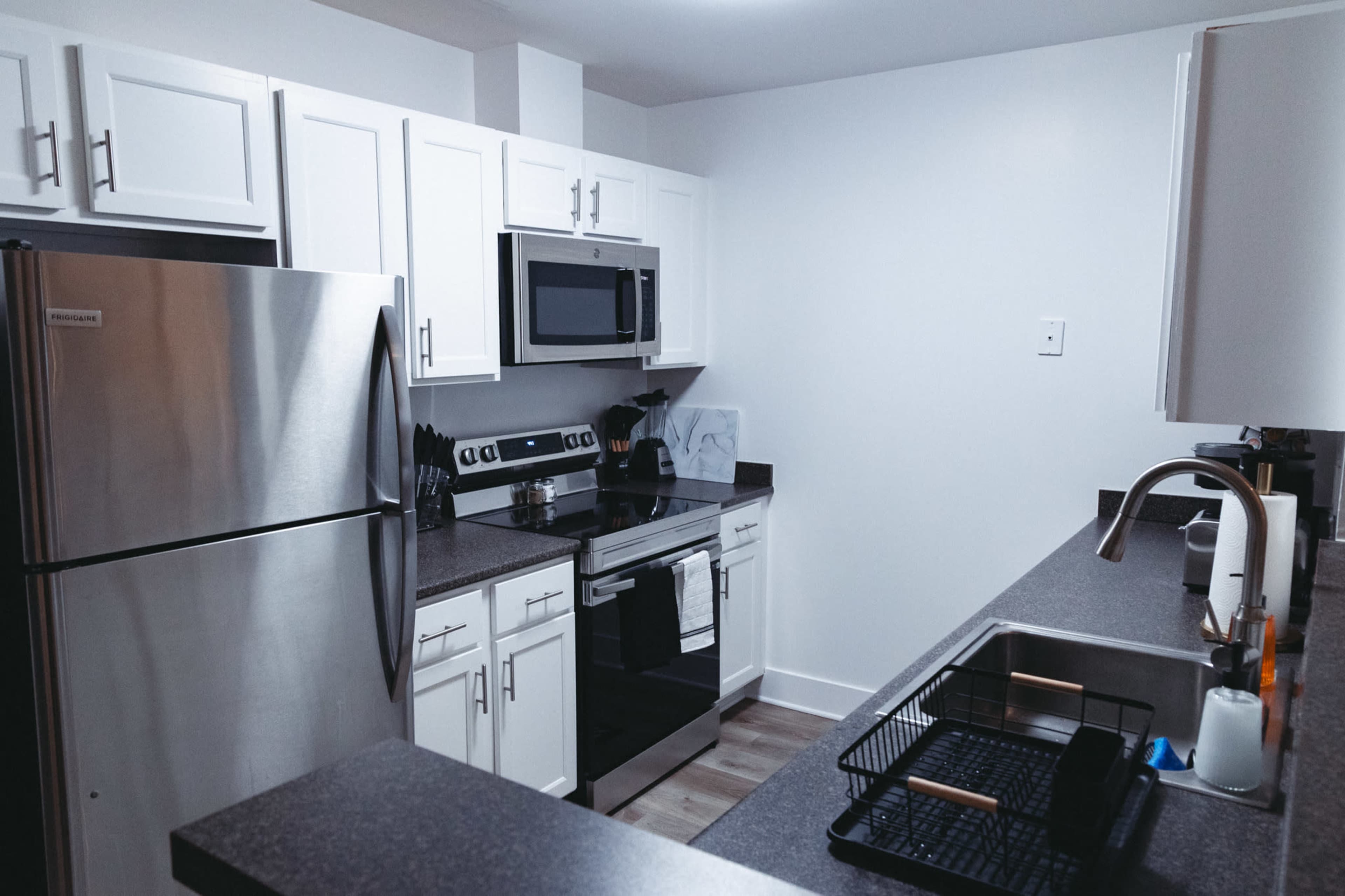 The image shows a modern kitchen with stainless steel appliances, white cabinetry, a dark countertop, and a dish rack by the sink.