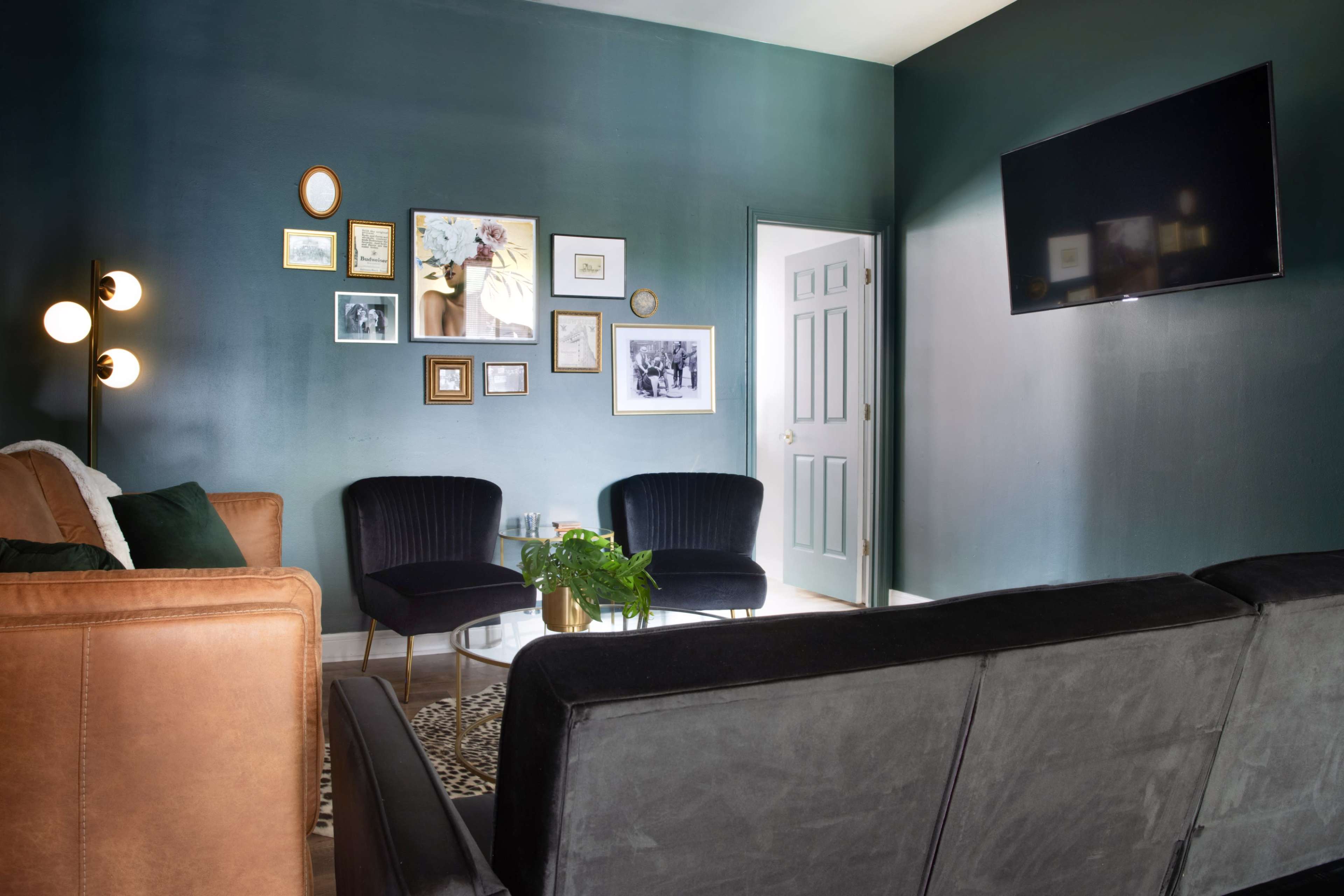 The living room features a dark green wall, a brown leather sofa, two black chairs, a glass coffee table, and a television mounted on the wall.