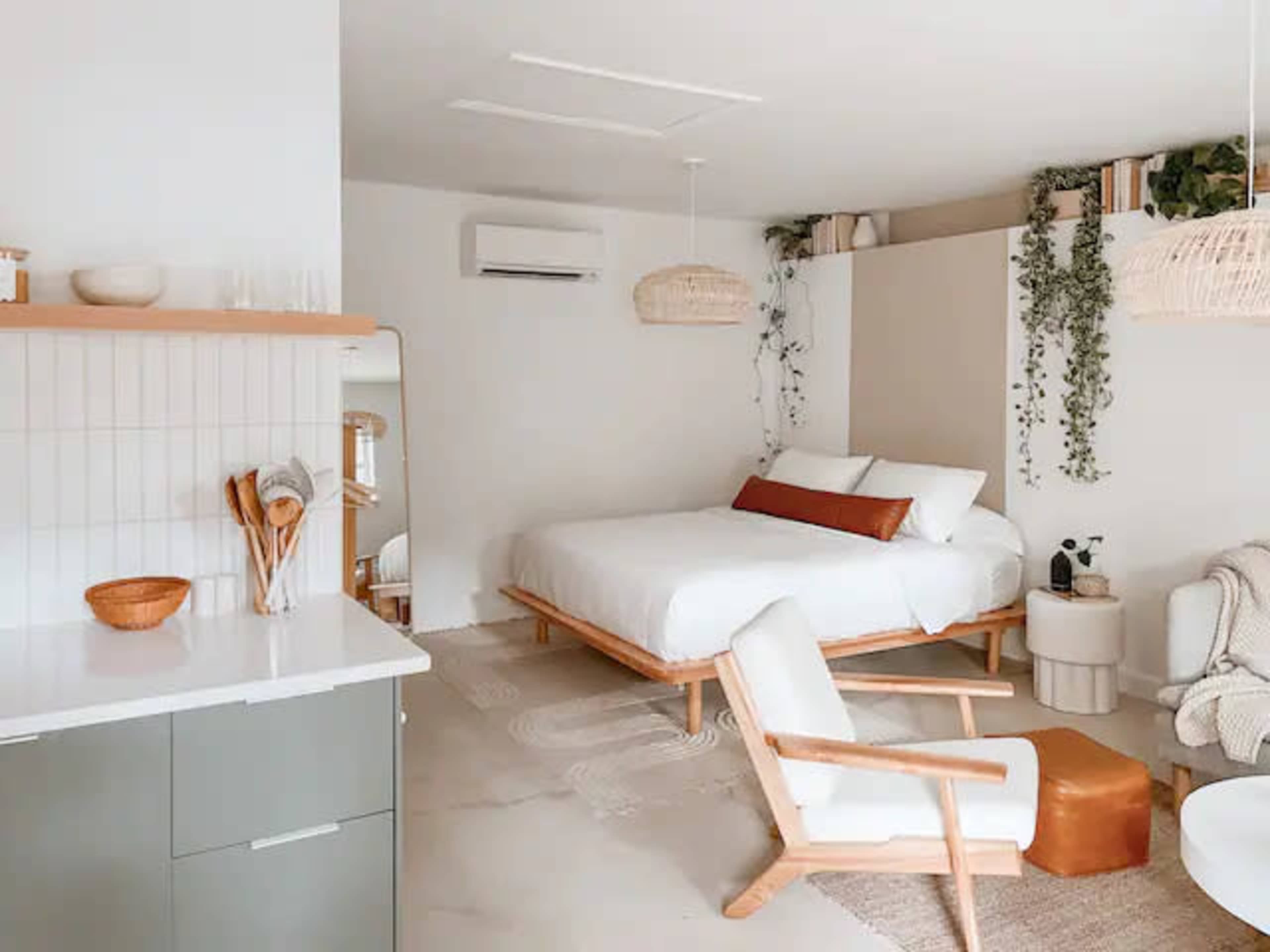 A modern, minimalist bedroom with a wooden bed, a small sitting area, and plants adorning the walls.