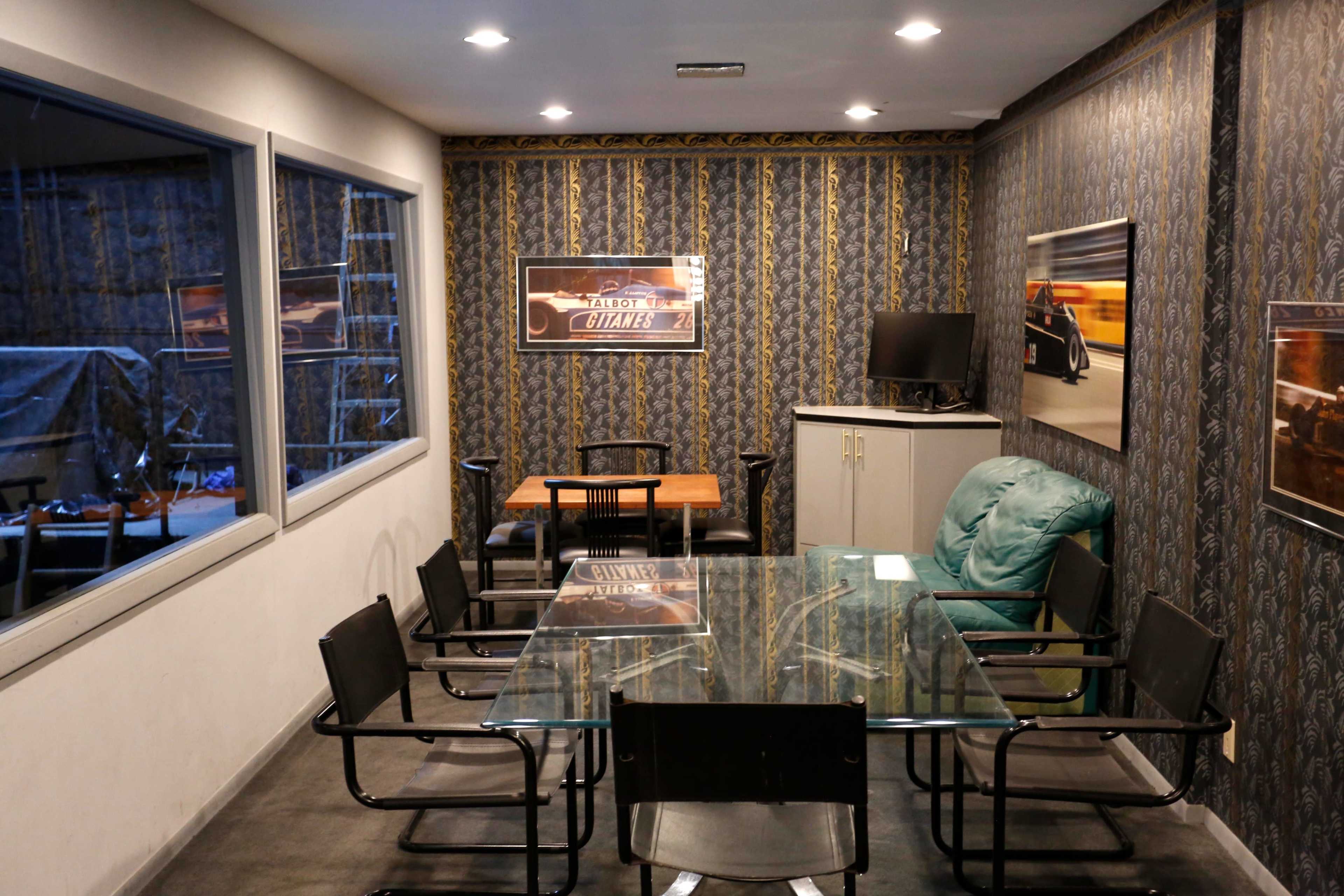 A small meeting room with a glass table, black chairs, a wooden table, a green armchair, and framed posters on the patterned walls.