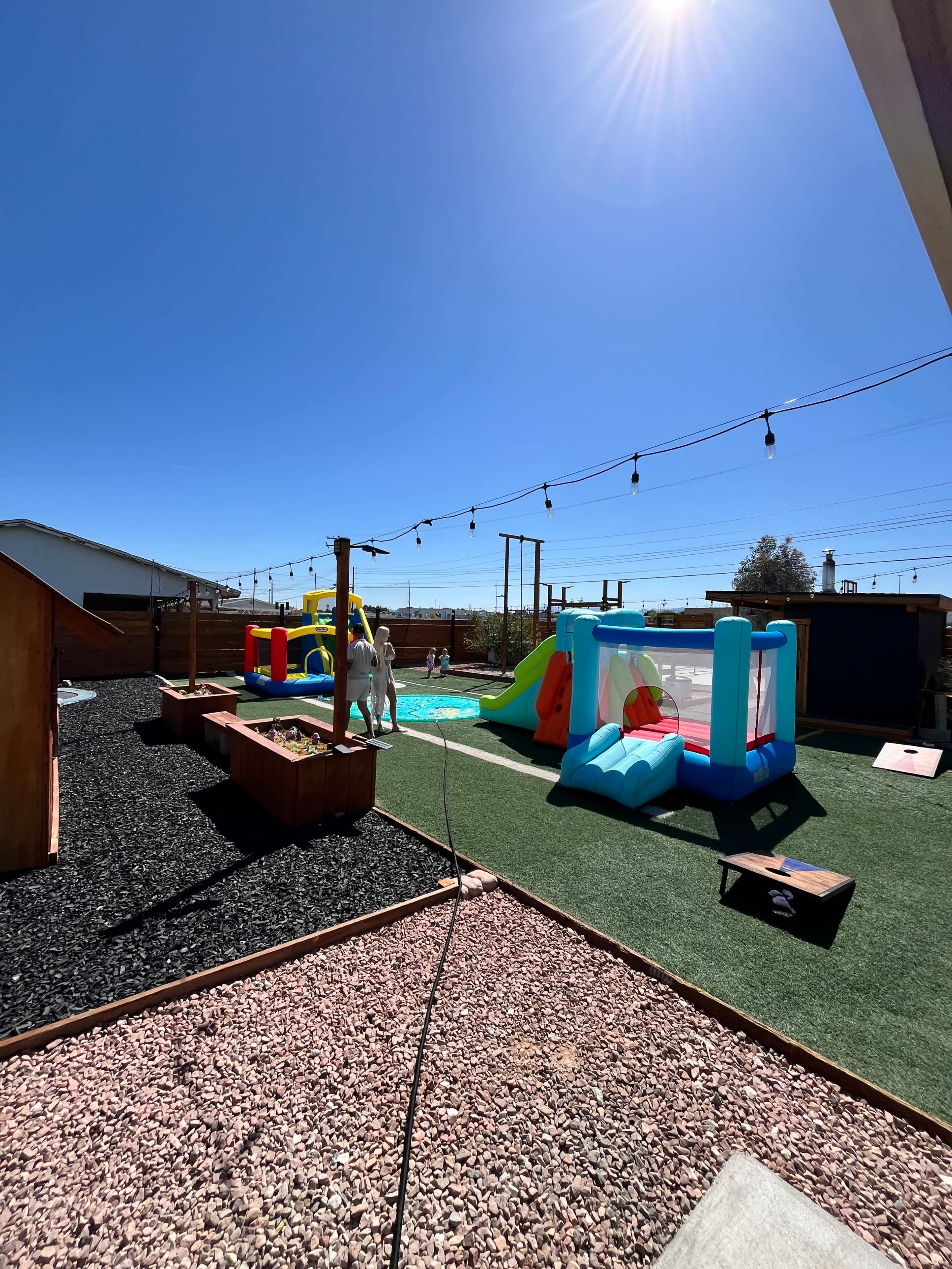 A backyard features a colorful inflatable play structure and a small water slide, surrounded by synthetic grass and a gravel pathway.