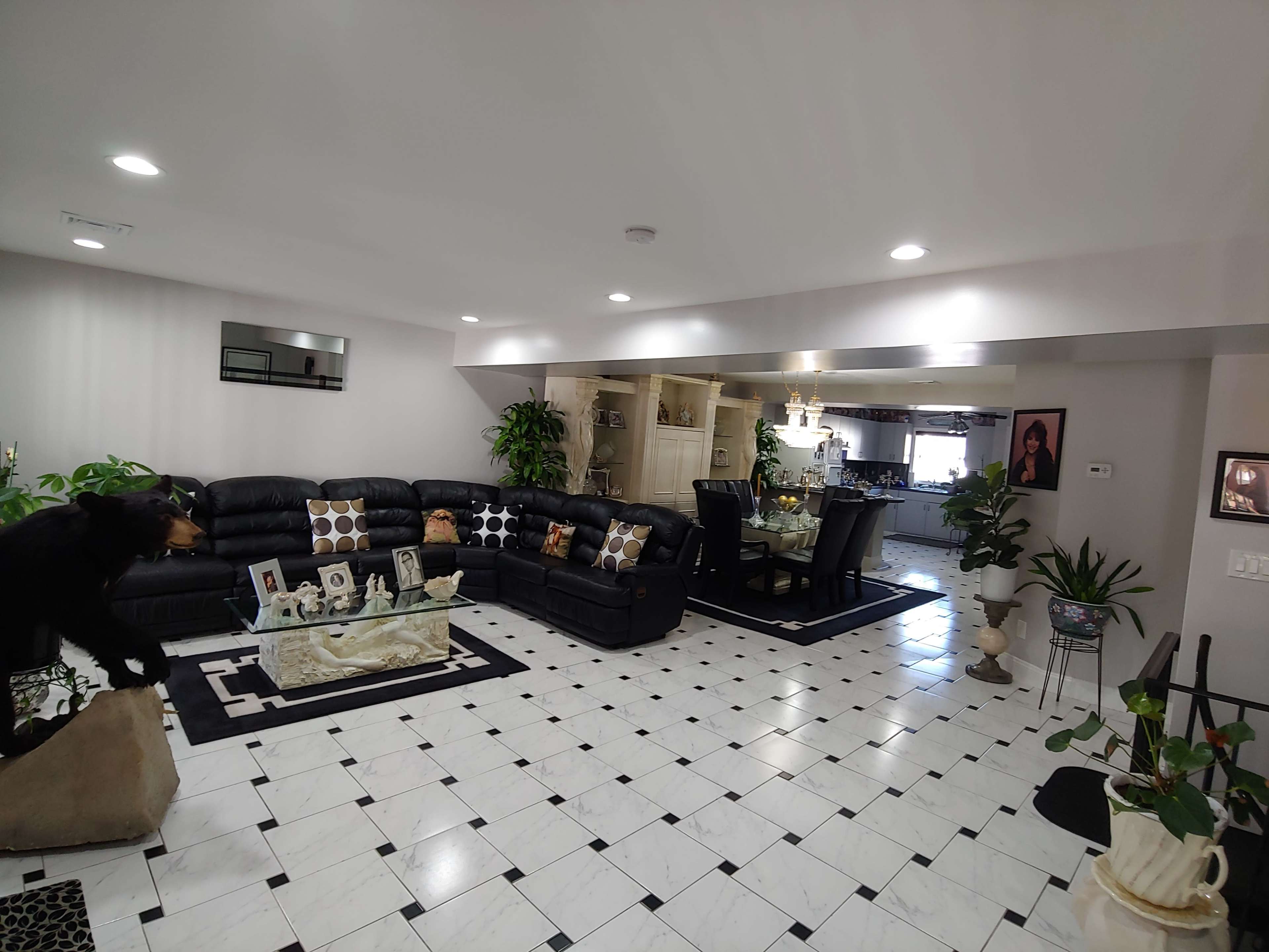 A living room with black leather furniture, a glass coffee table, and a dining area in the background, featuring a tiled floor with black and white patterns.