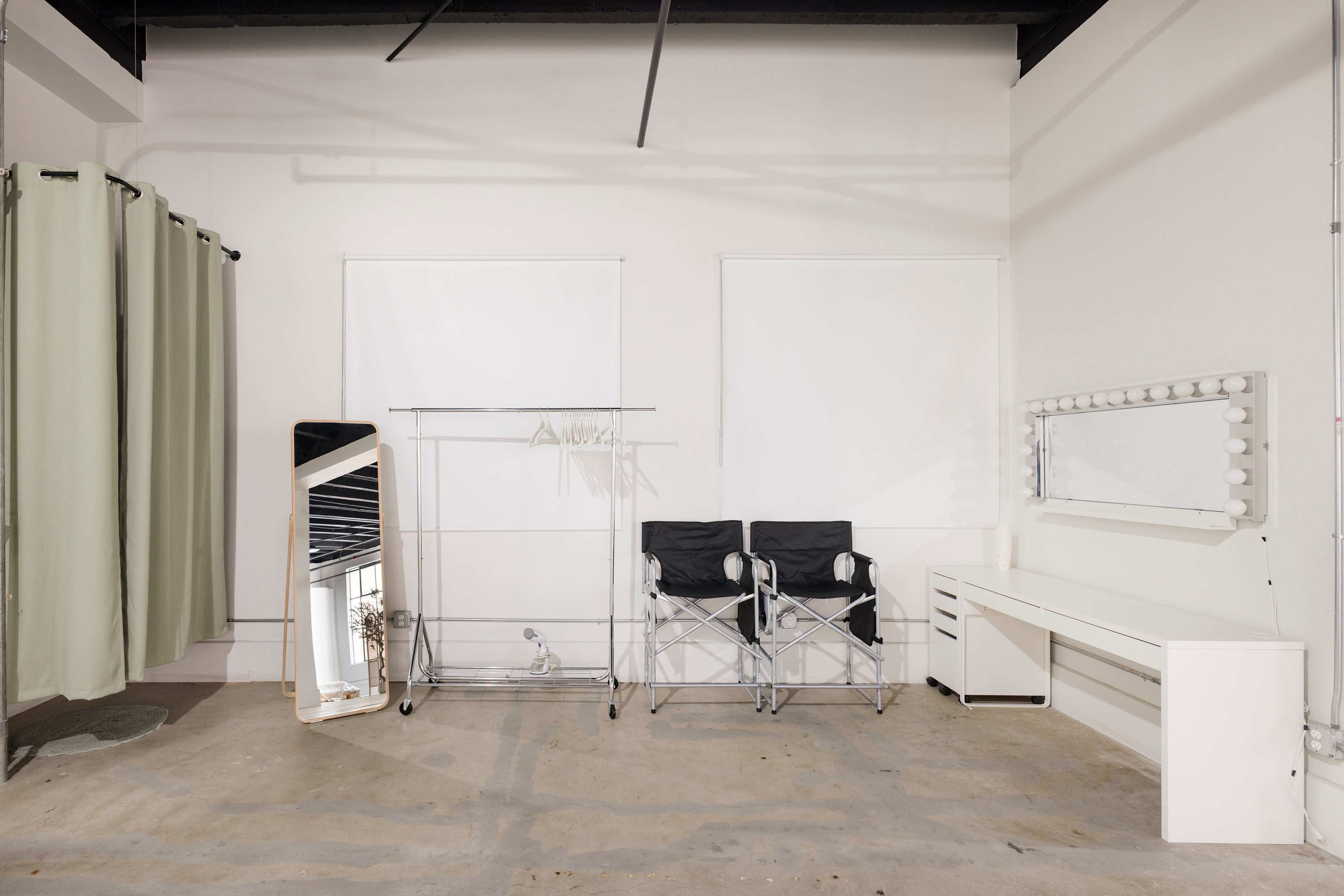 The image shows a minimalist dressing room with two director's chairs, a large mirror with lights, a rack for clothes, and green curtains along the walls.