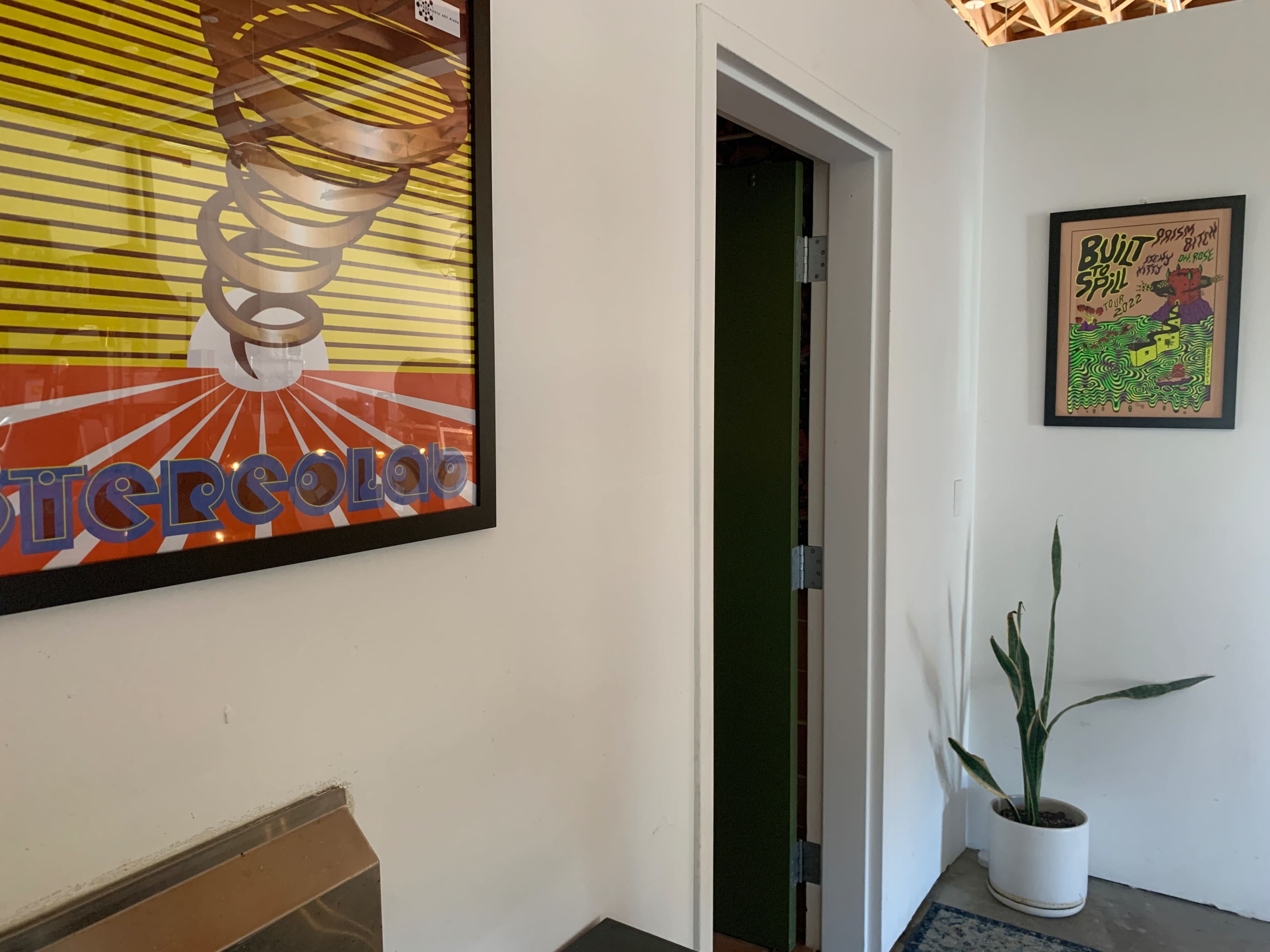 The image shows a well-lit interior space with framed artwork on the walls, a door leading to another room, and a potted plant in the corner.