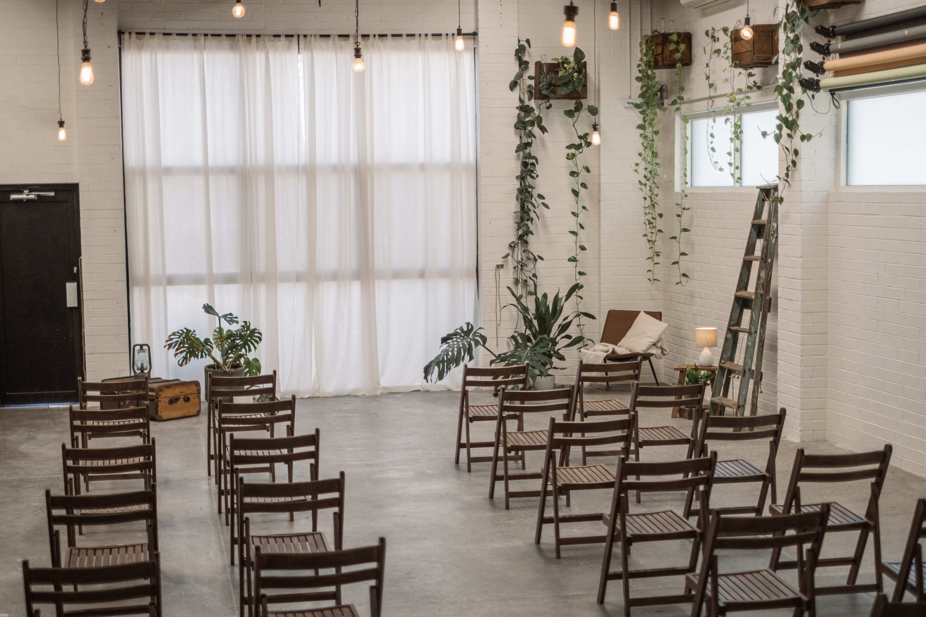 A spacious indoor venue with wooden chairs arranged for an event and tall white curtains, accented by plants and light fixtures.