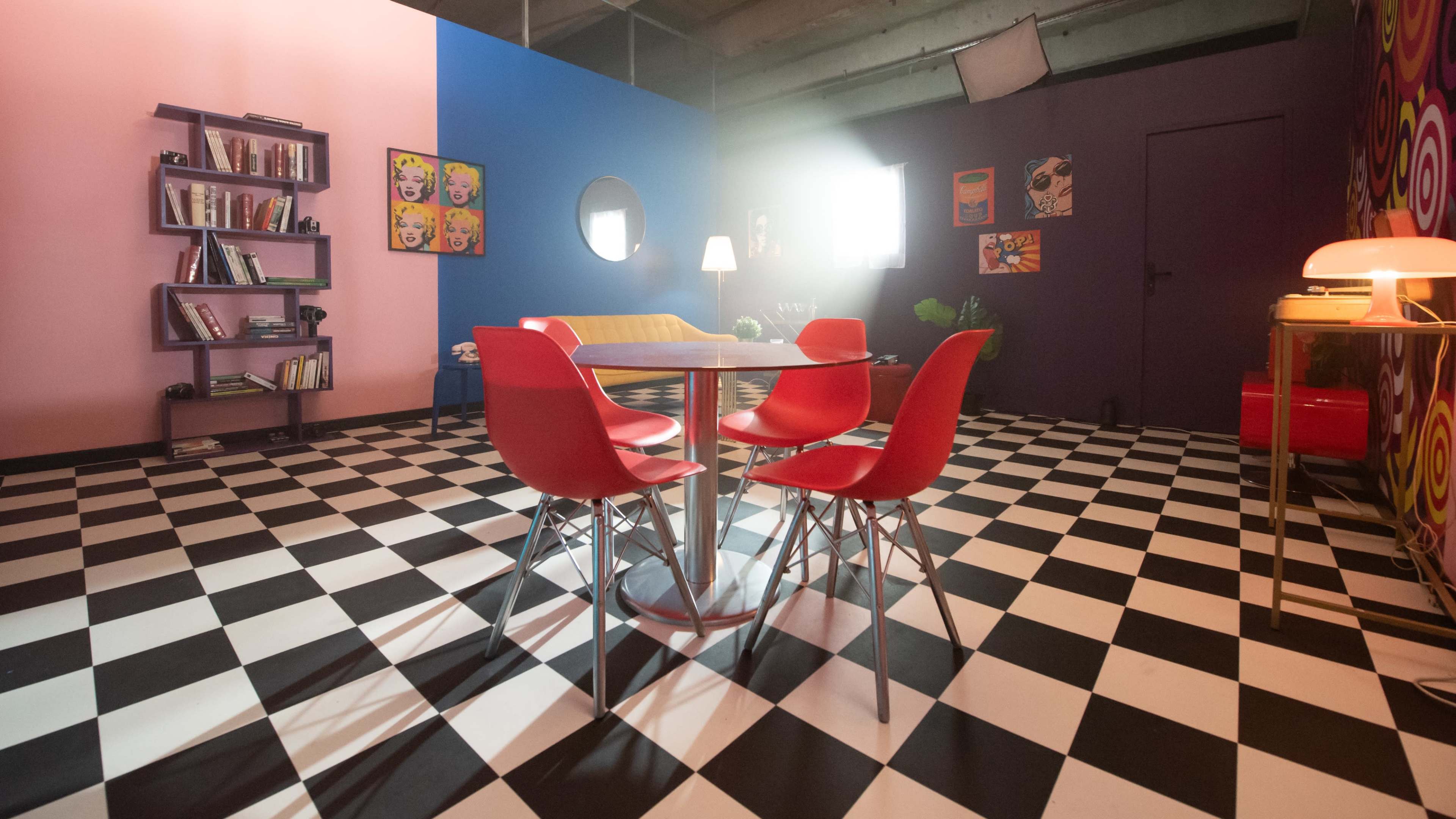 A modern room with a checkered floor, colorful walls, a circular mirror, and a dining table surrounded by four red chairs.