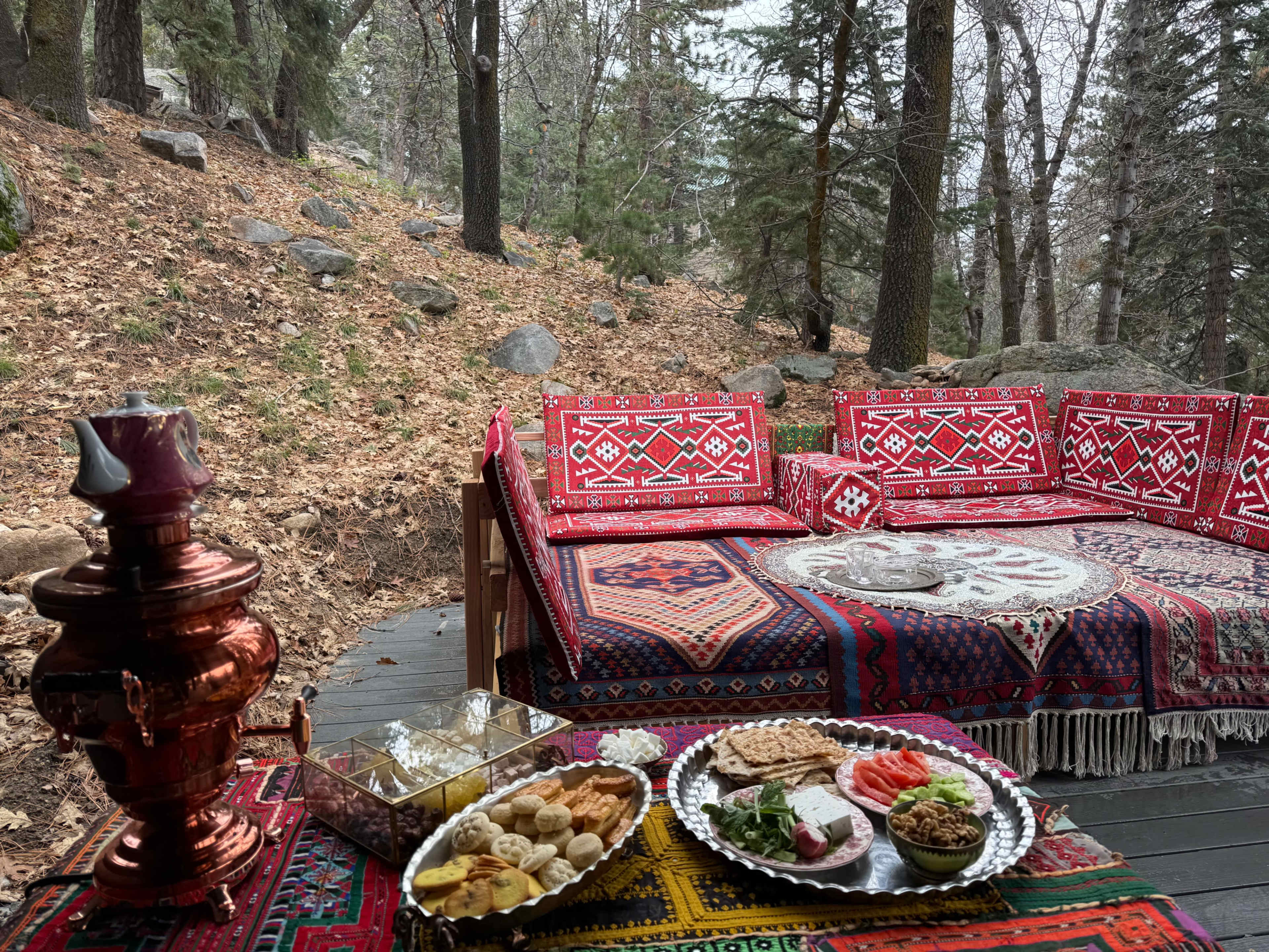 The scene features a cozy outdoor seating area adorned with traditional rugs, alongside a table set with a variety of dishes and a decorative samovar, surrounded by a forested backdrop.