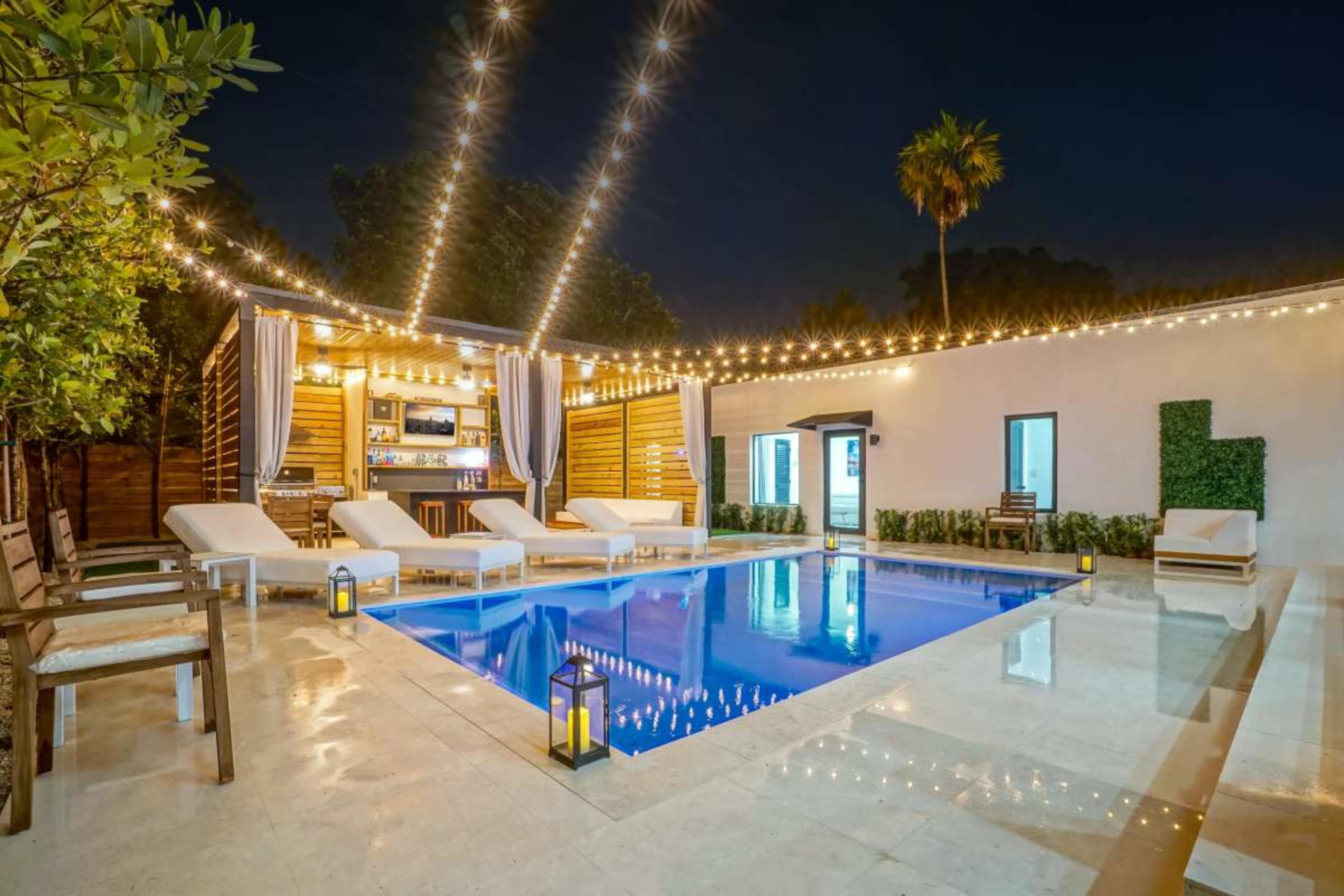 The image shows a modern outdoor space featuring a swimming pool, lounge chairs, and a bar area, illuminated by string lights at night.