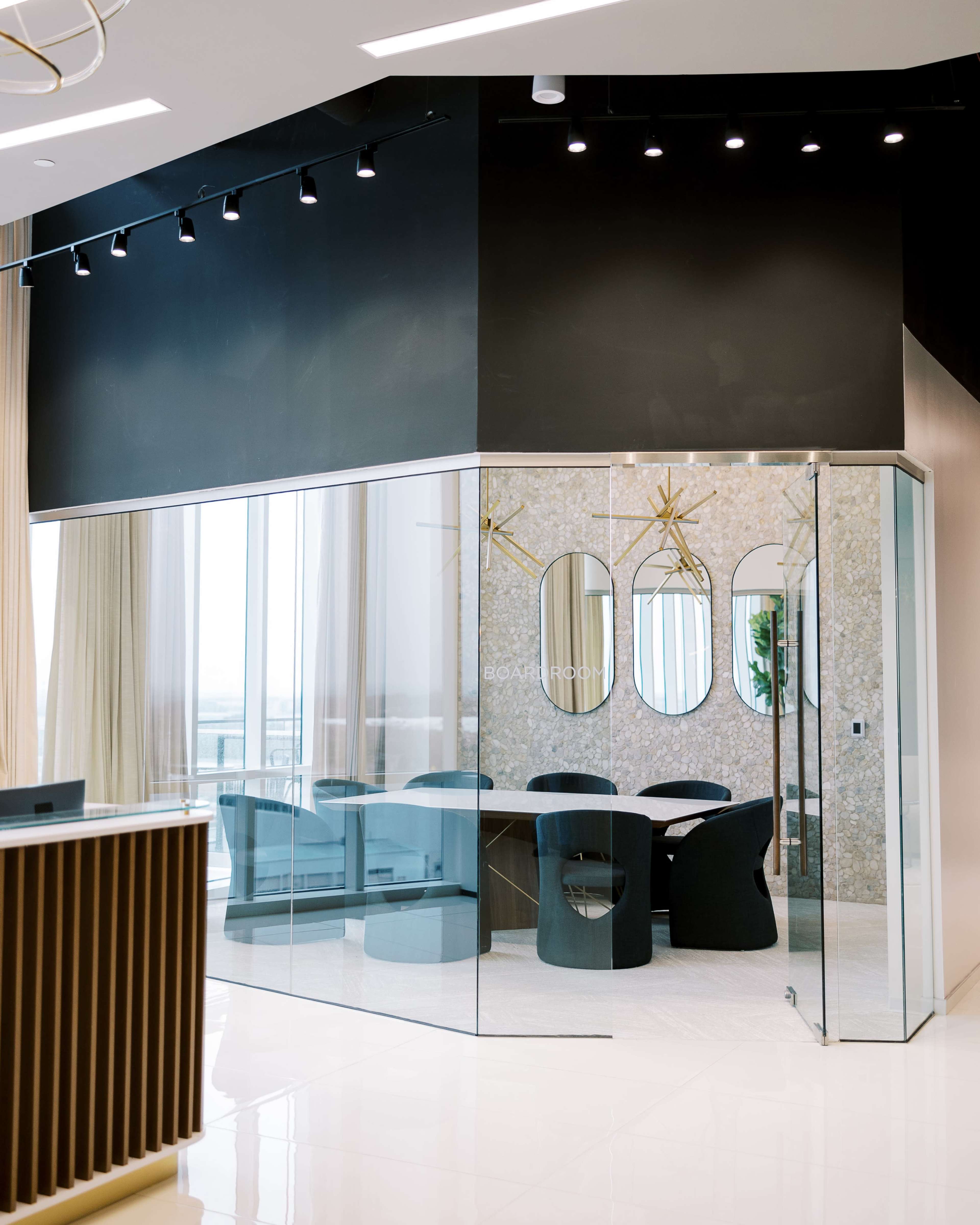 The image shows a modern conference room enclosed in glass, featuring a large table and black chairs, with three round mirrors on the wall and large windows providing natural light.