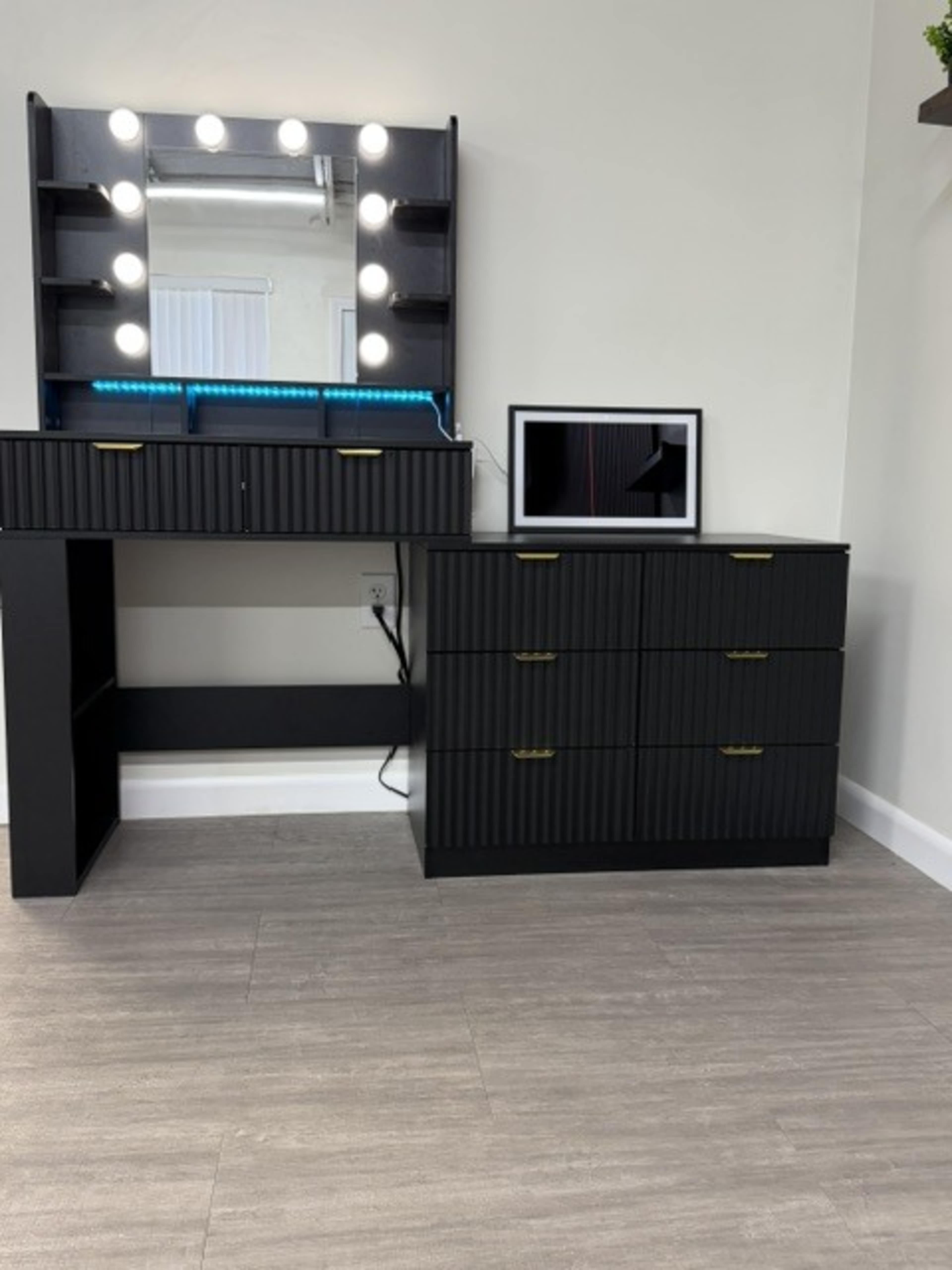 The image shows a modern makeup vanity with bright lights on top and a matching dresser with multiple drawers beside it, all set against a light-colored wall.
