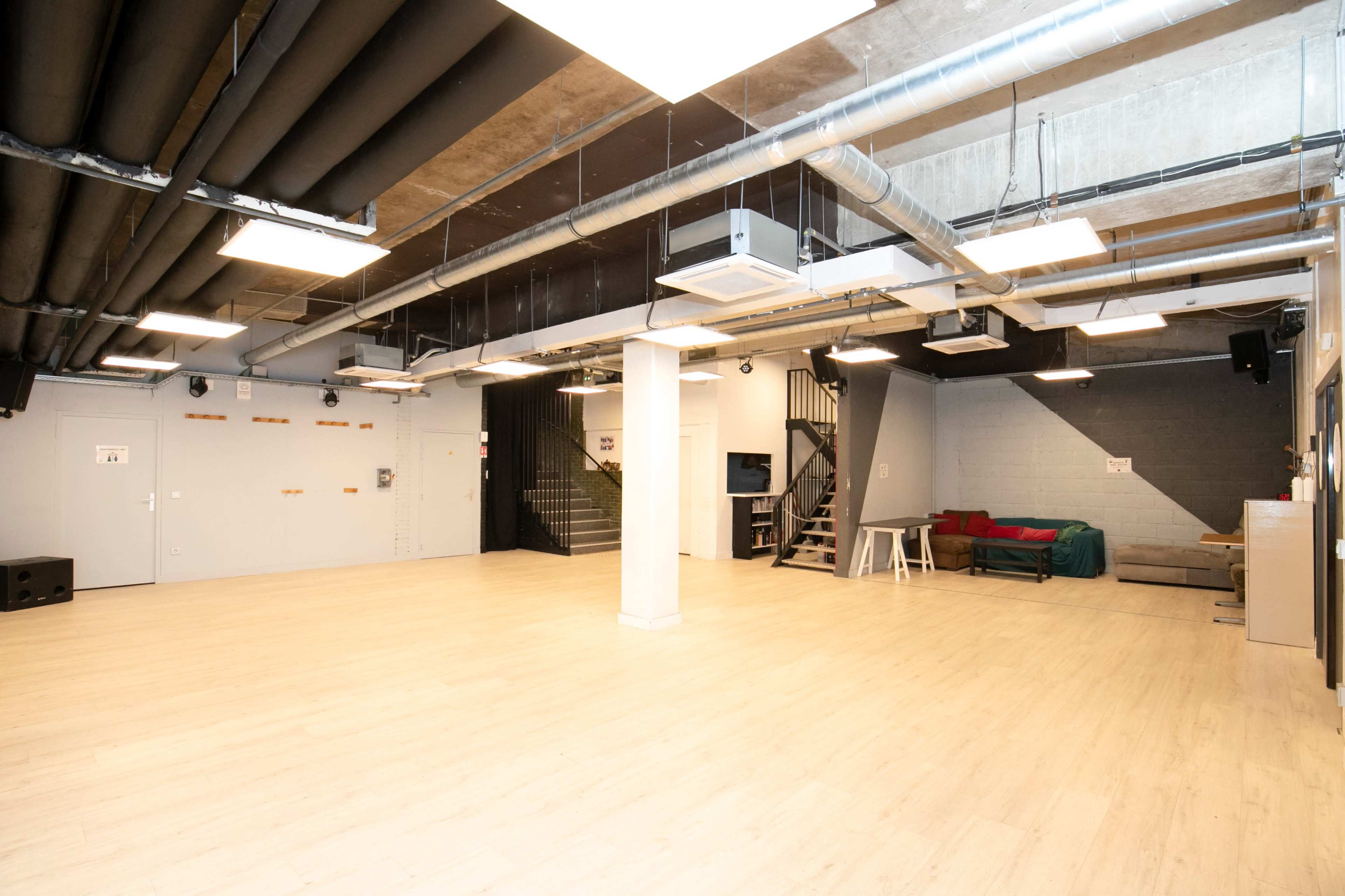 A spacious, empty room with light wooden flooring and industrial-style ceilings, featuring a staircase in the back and a small couch against the wall.